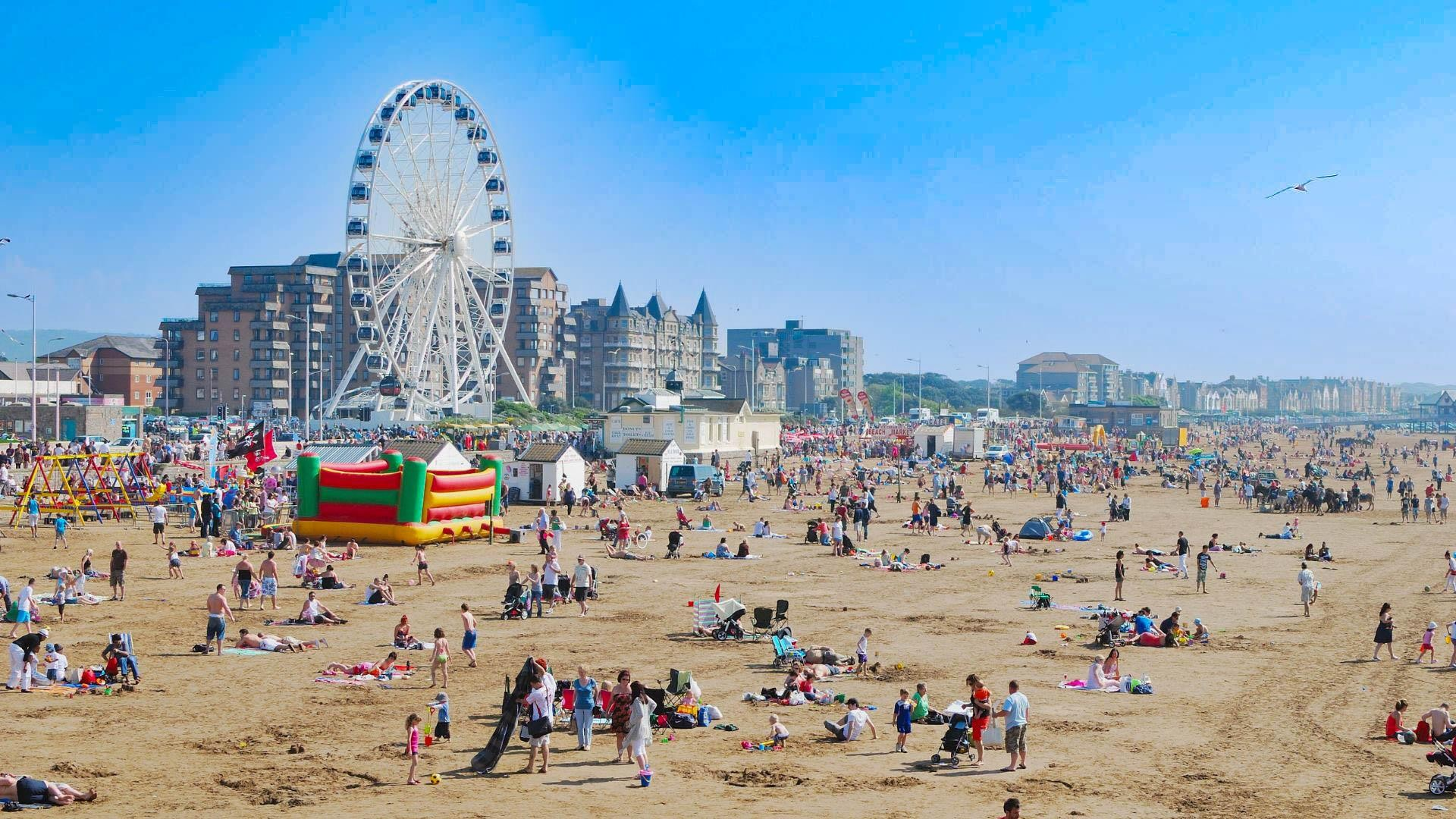 Crowded beach scene with many people relaxing and playing, amusement park ferris wheel, high-rise buildings in the background, blue sky with a few seagulls.