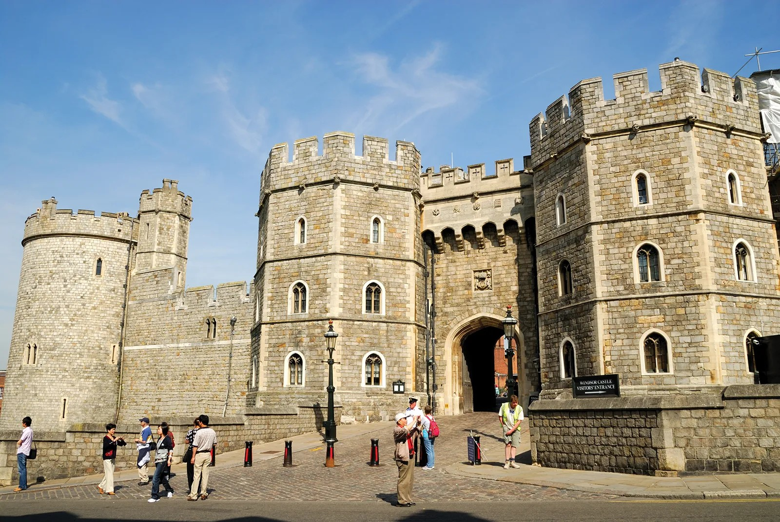 Windsor Castle's entrance with stone walls, towers, and a large archway, with visitors and a guard outside on a sunny day.
