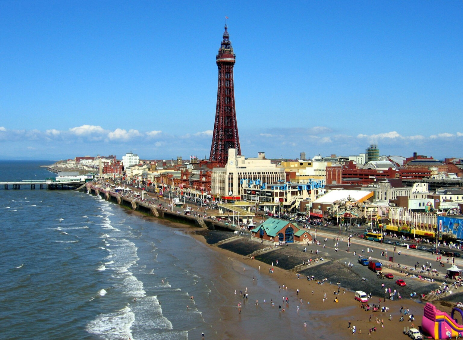 View of the coastline and amusement park with the iconic Blackpool Tower, blue sky, and people on the beach and promenade.