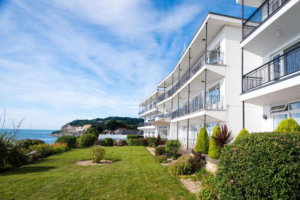 White seaside apartment building with balconies overlooking a green lawn, bushes, and the ocean under a blue sky