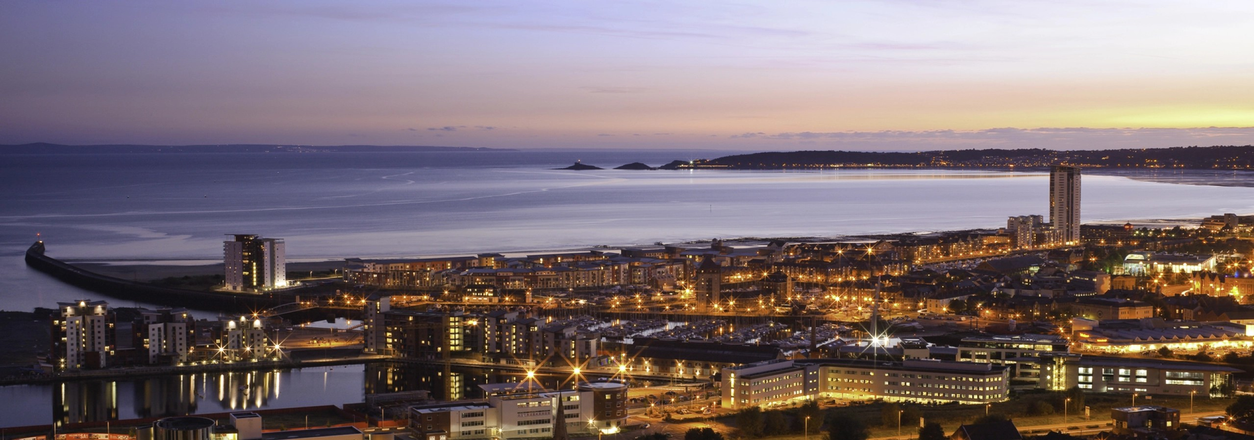 A cityscape at dusk with buildings illuminated, a calm body of water reflecting the lights, and a sky transitioning from sunset to night.