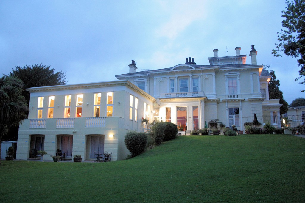 A large, elegant white mansion with lit windows and a well-maintained green lawn at dusk.