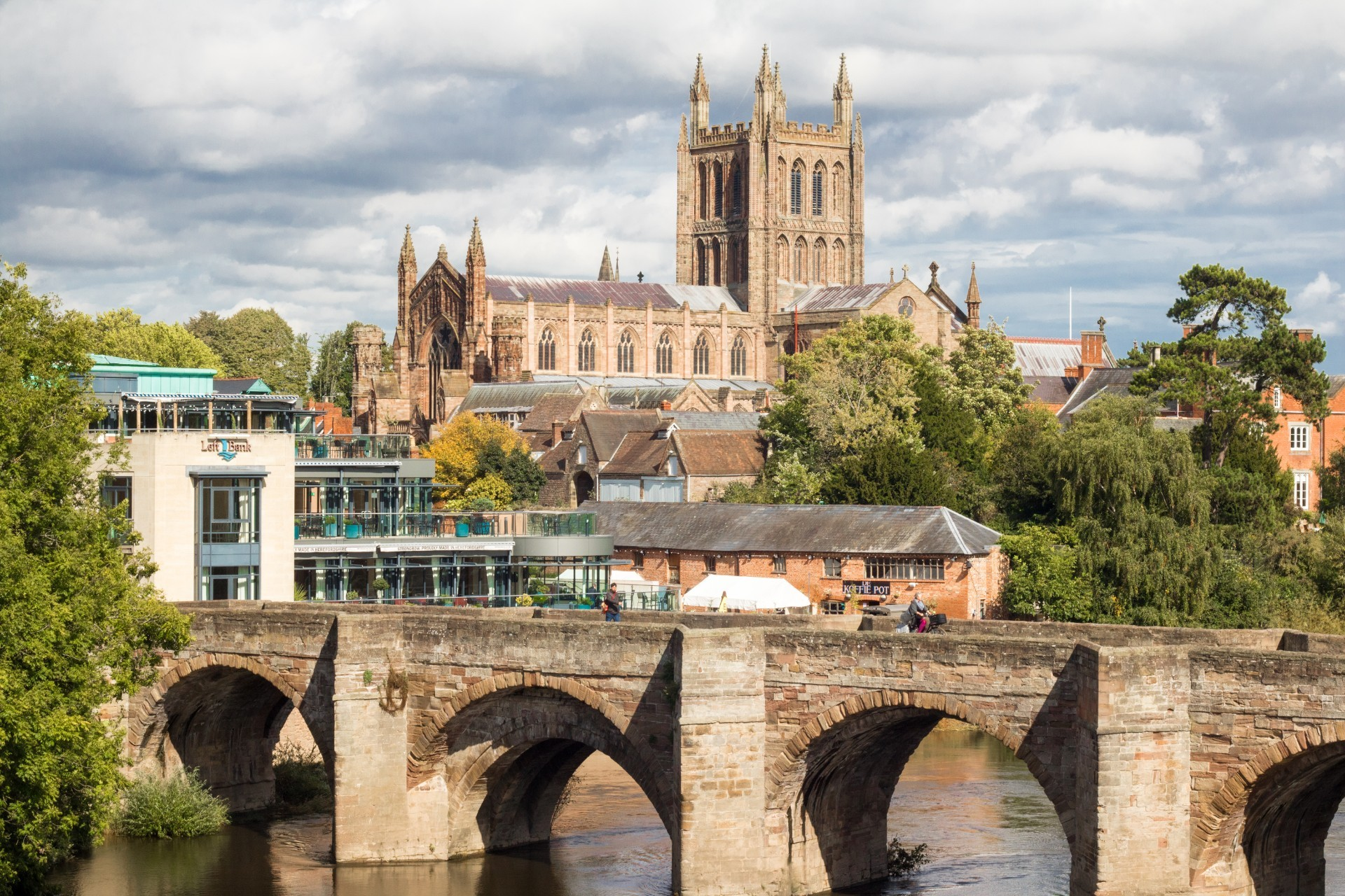 A scenic view of a historic stone bridge over a river with trees on either side. In the background, a large Gothic-style church with tall spires and stained-glass windows rises above the surrounding buildings. Modern buildings and people walking on the bridge are also visible.