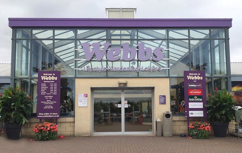 Exterior of a garden center named Webbs, with large glass windows and purple signage, adorned with potted plants and flowers outside.