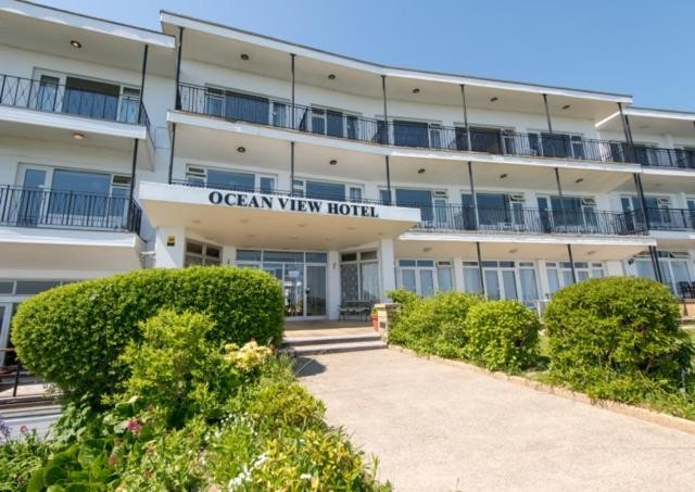 Front view of Ocean View Hotel with bushes and a walkway leading to the entrance, multiple balconies on each floor, and a clear blue sky.