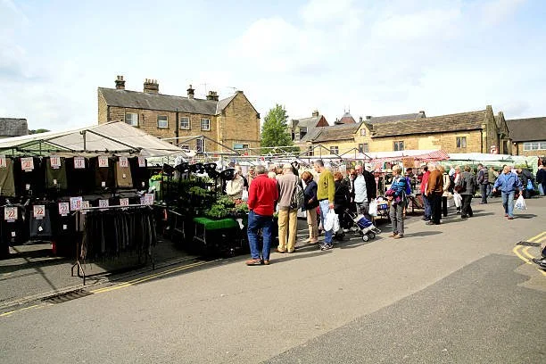 People shopping at an outdoor market stall in a small town parking lot with houses in the background.