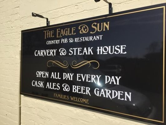 Black signboard with gold and white lettering advertising The Eagle & Sun, a country pub and restaurant, claiming it is a carvery and steak house, open daily all day, with a cask ales and beer garden, welcoming families.
