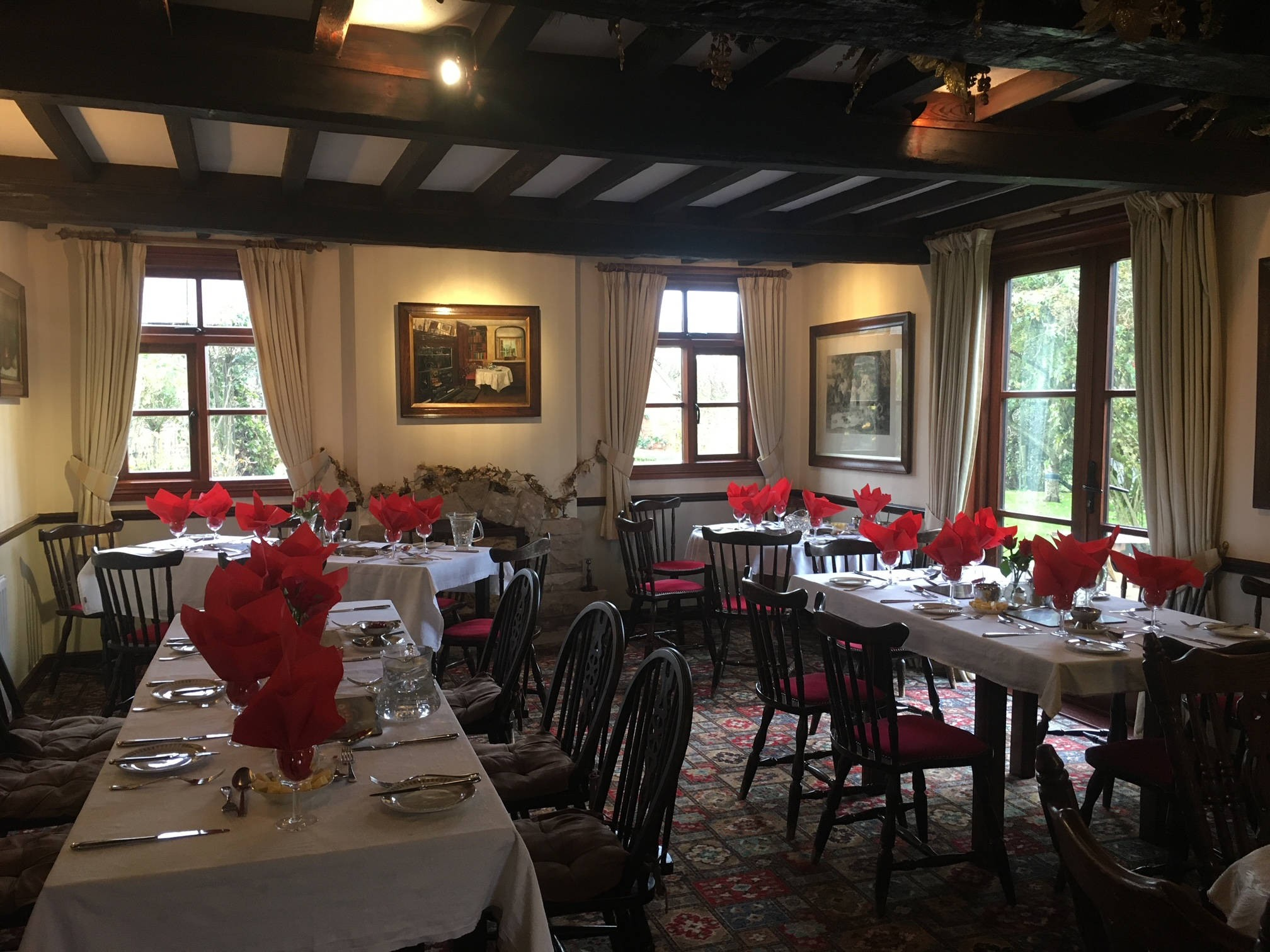 Finely set dining room with multiple tables covered in white tablecloths, decorated with red napkins folded in a floral shape, glassware, and cutlery. The room has large windows with cream curtains, traditional paintings on the walls, and a patterned rug on the floor.