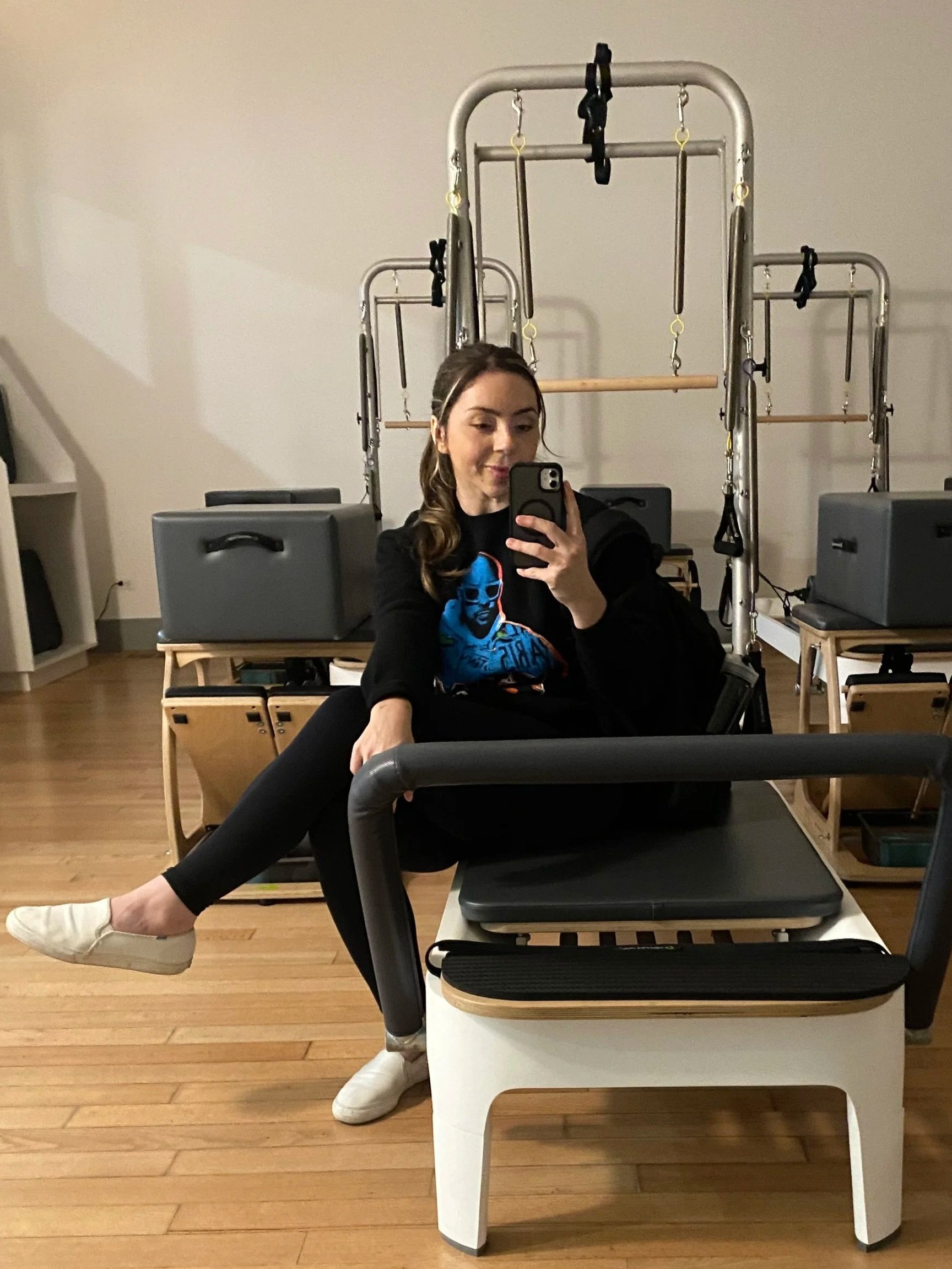 A woman taking a selfie in a fitness or physical therapy studio with Pilates or exercise equipment in the background.