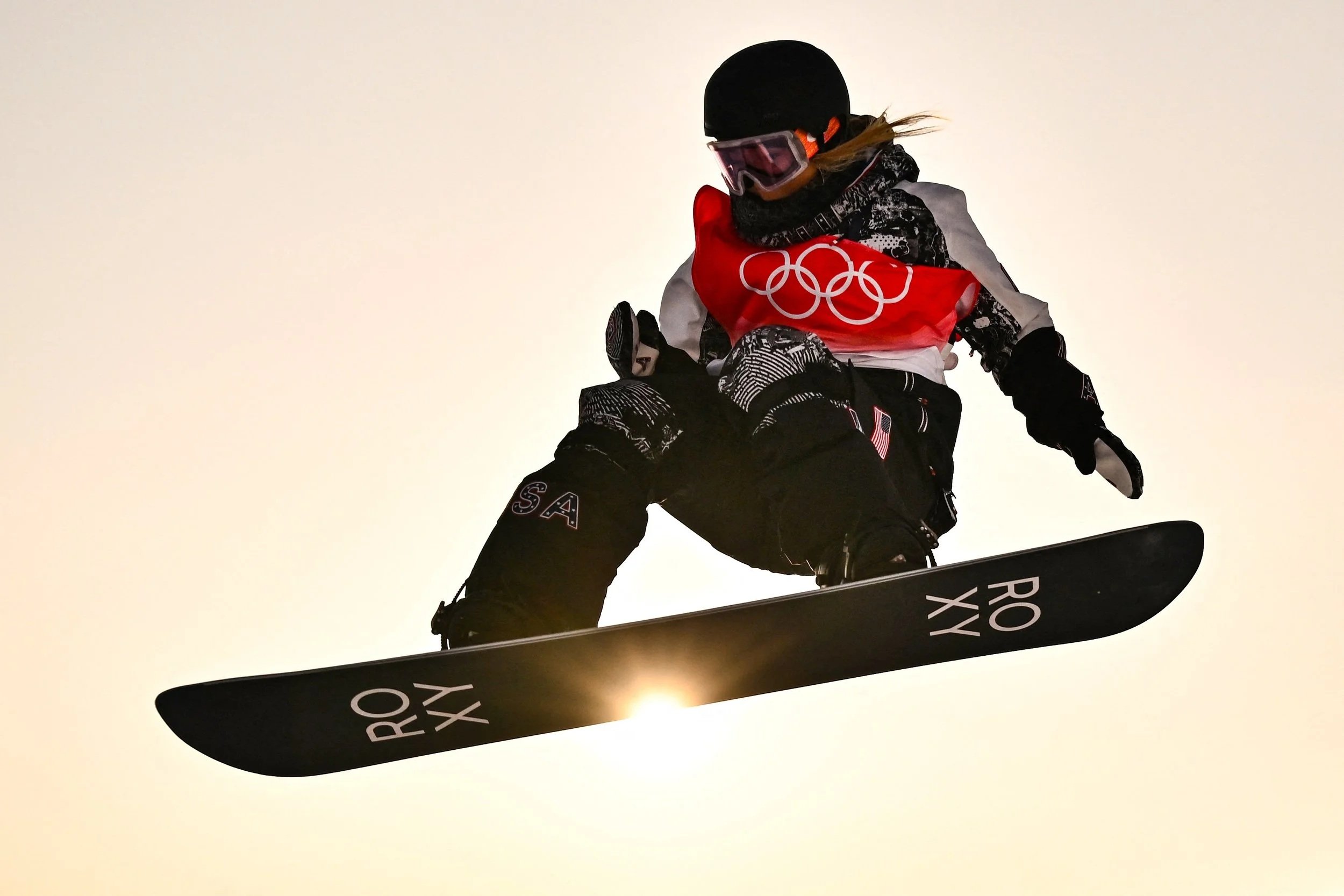 Chloe Kim mid-air during an Olympic halfpipe run, grabbing her snowboard against a golden sky