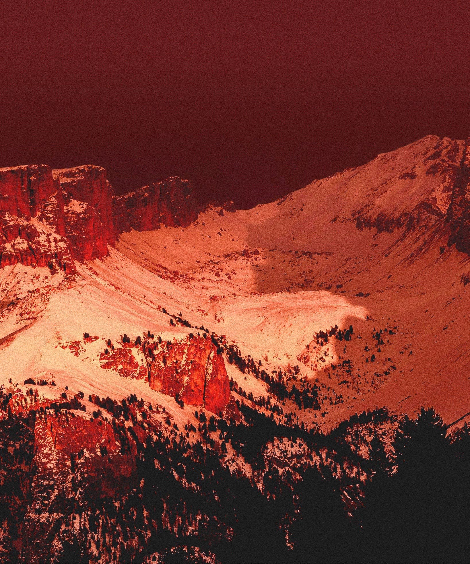 Snow-covered mountain peaks and forested slopes under a deep red sky