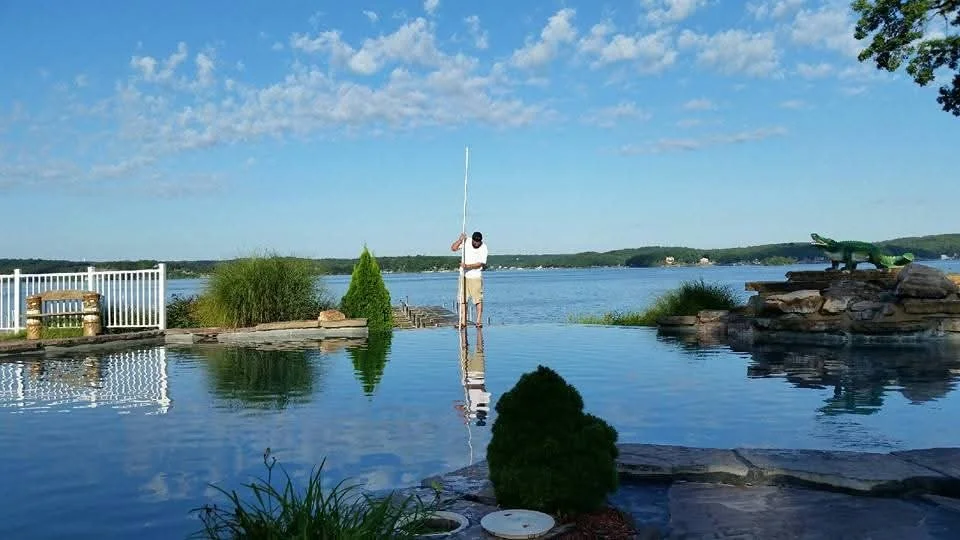 A man in white shirt and shorts fishing in a backyard pool with a view of a lake and blue sky in the background.