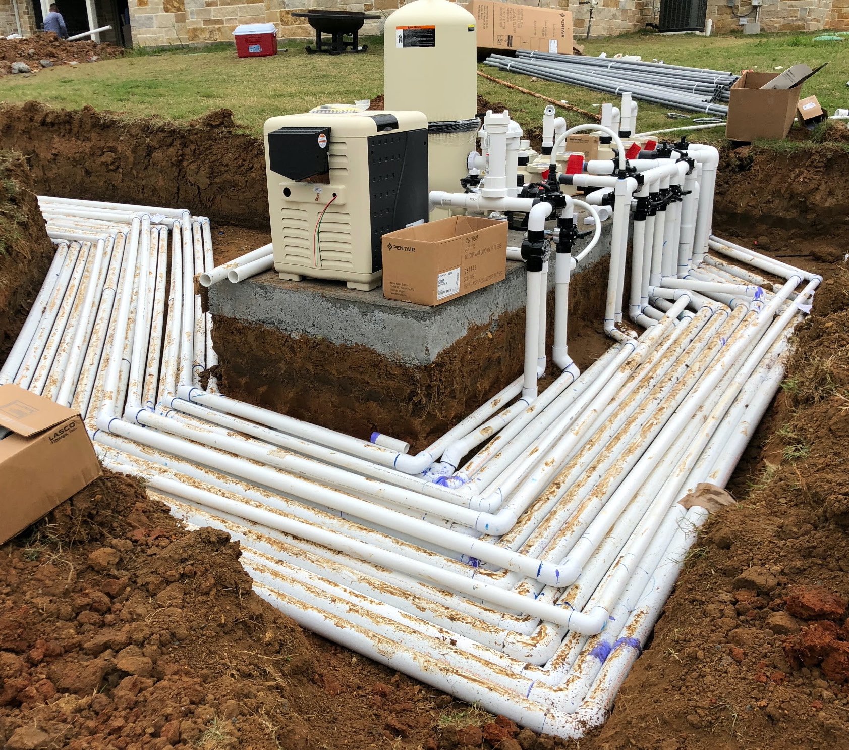 Underground plumbing system with white pipes, a water filtration or pump unit, and other equipment on a concrete slab at a construction site.