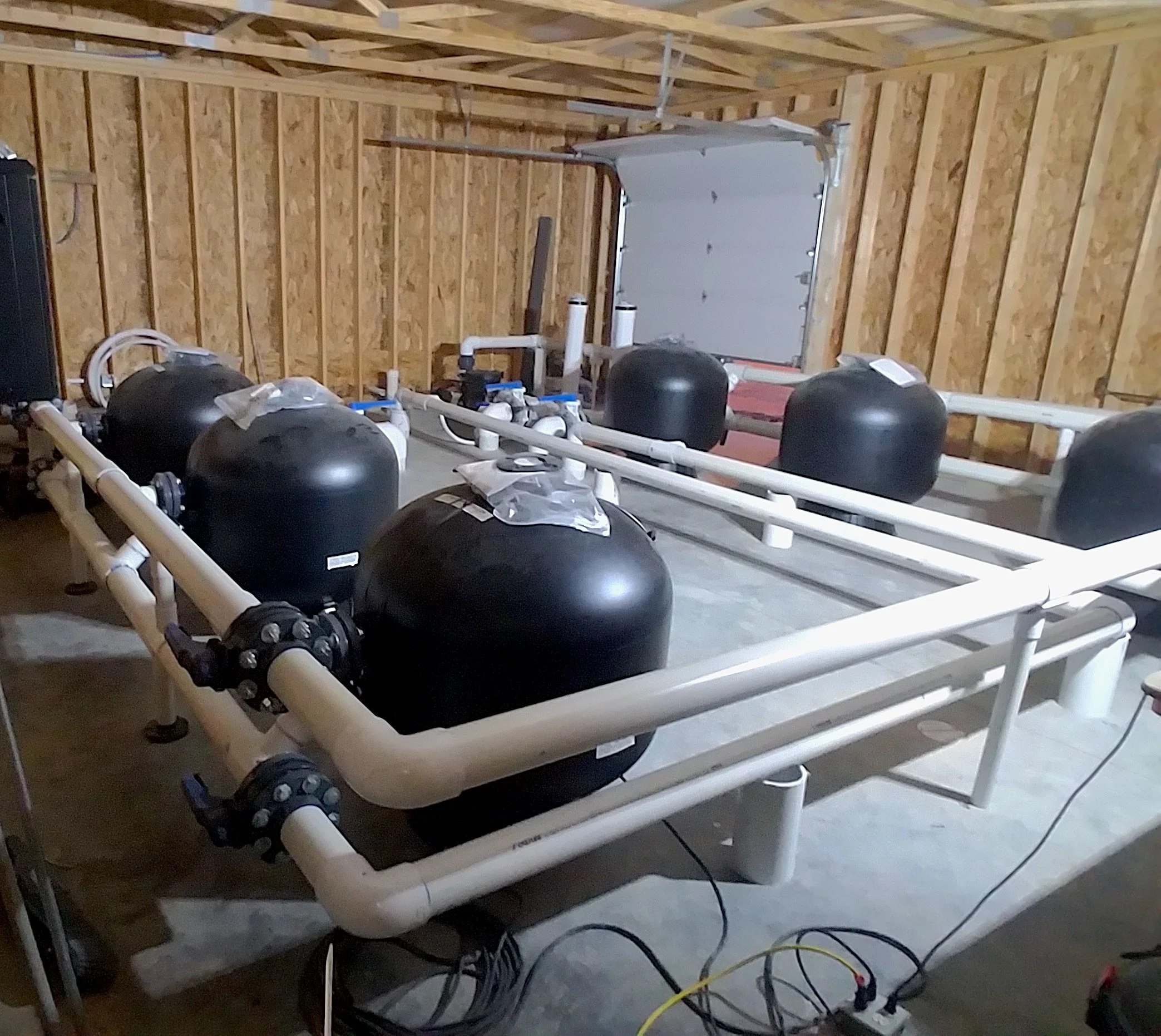 A construction site of a home pool with black water filters connected to white PVC pipes on a concrete floor in a wooden-sided garage or workshop space.