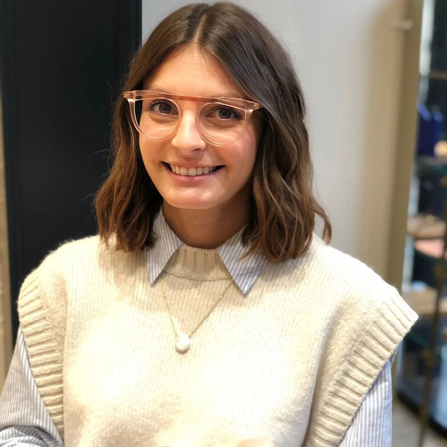 Jeune femme souriante portant des lunettes roses, pull beige, collier et chemise à rayures, en intérieur.