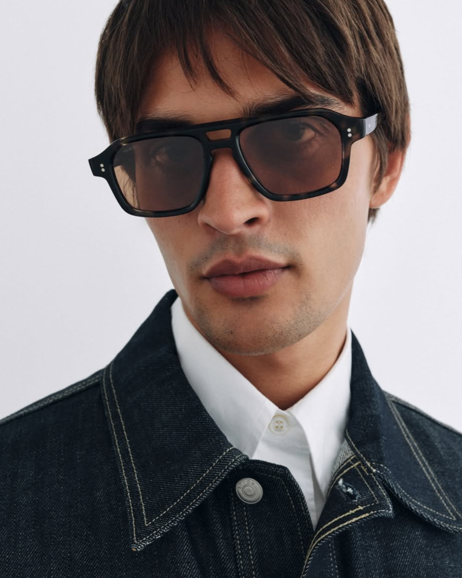 Homme portant des lunettes de soleil foncées et une veste en denim, avec une chemise blanche, visage proches de l'objectif, fond blanc.