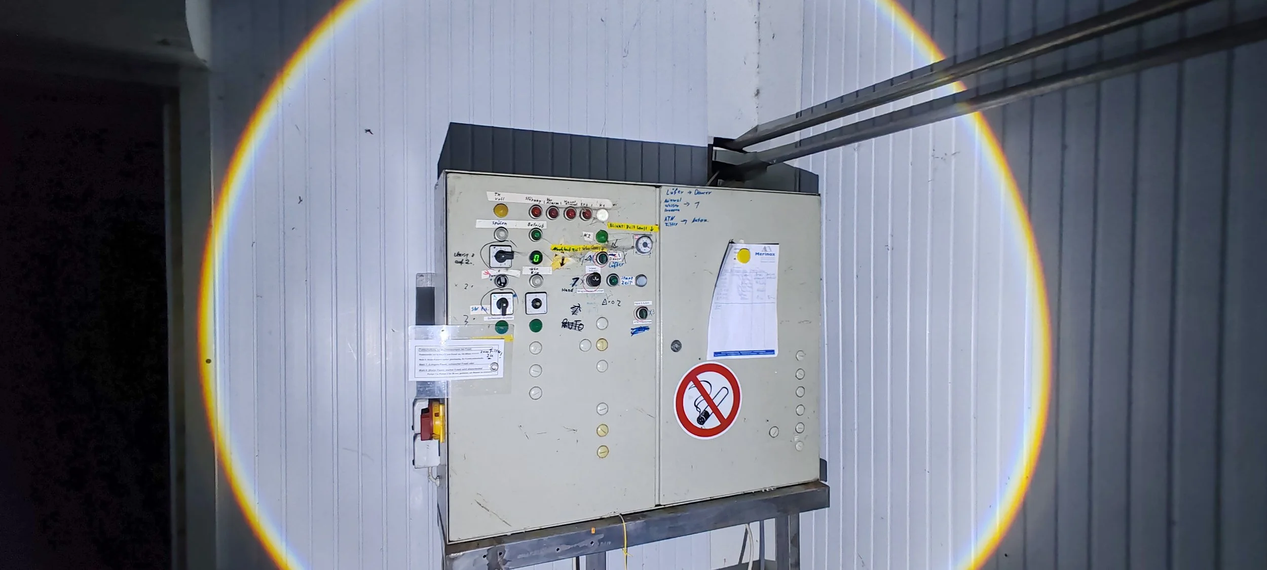 Schaltkasten an einer Wand mit mehreren Schaltern, Kontrollleuchten, Not-Aus-Schalter, Notizzetteln und einem Verbotsschild, das das Rauchen verbietet.