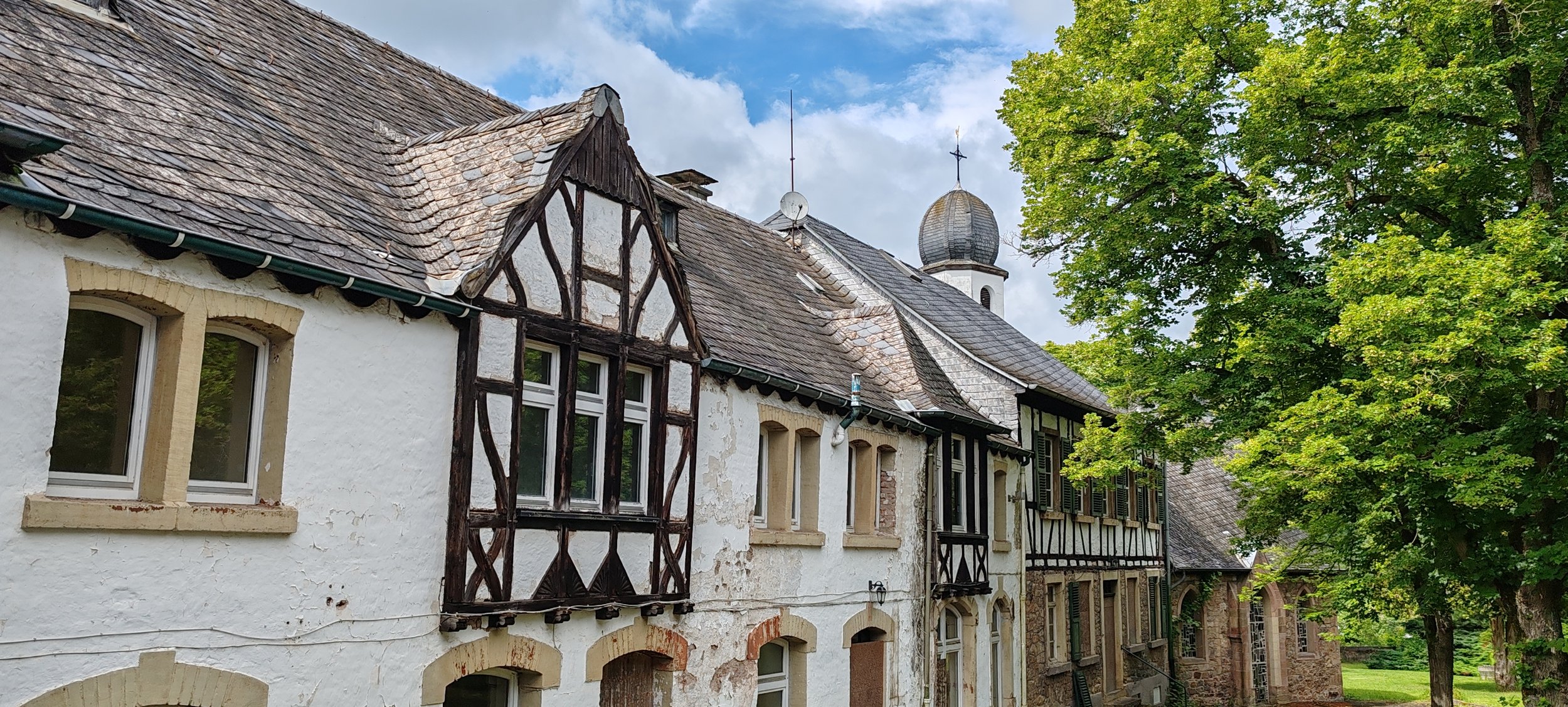 Historisches Gebäude mit Fachwerk- und Mauerfassade, umgeben von großen, grünen Bäumen, mit einem Himmel mit Wolken im Hintergrund.