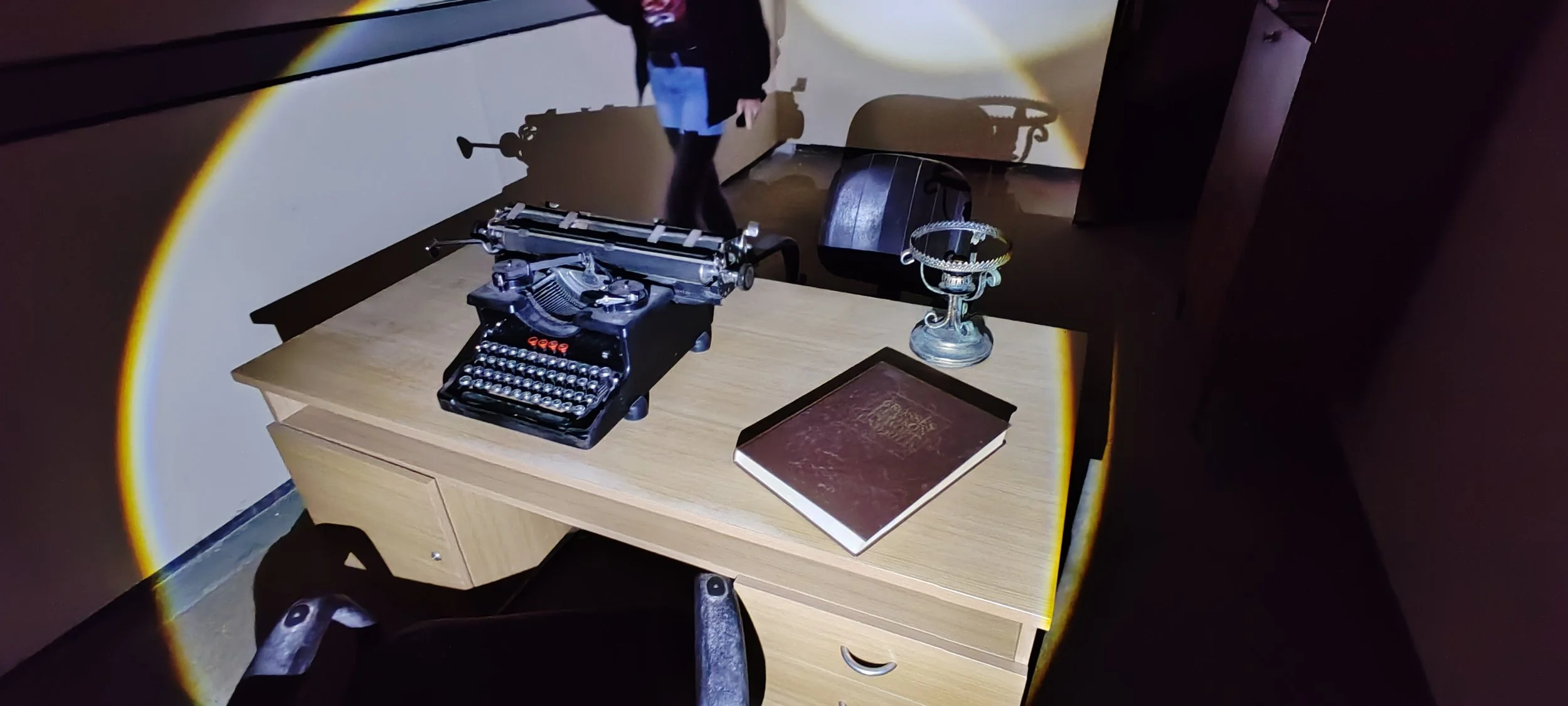 Ein Schreibtisch mit einer alten Schreibmaschine, einem Buch, einer Trophäe und einem Laptop. Im Hintergrund eine Wand mit Projektionsbildschirm, auf dem eine Person zu sehen ist.