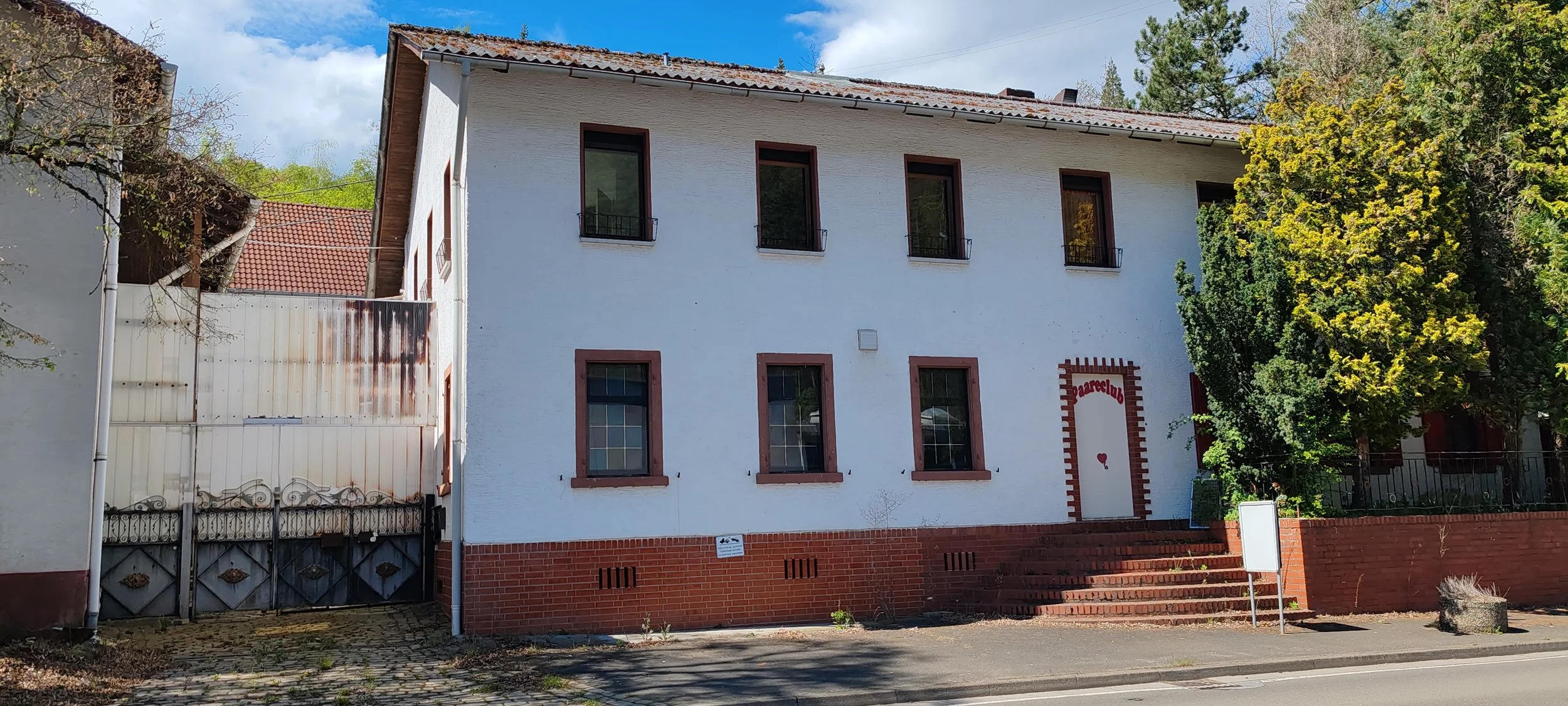 Hauptgebäude mit weißer Fassade, vier Stockwerke, rote Fensterrahmen, rote Ziegelmauer im unteren Bereich, Eingang mit rot-weißer Umrandung und Herzsymbol, Stufen vor Eingang, umgeben von Bäumen und Hecken, Straße und Gehweg im Vordergrund.