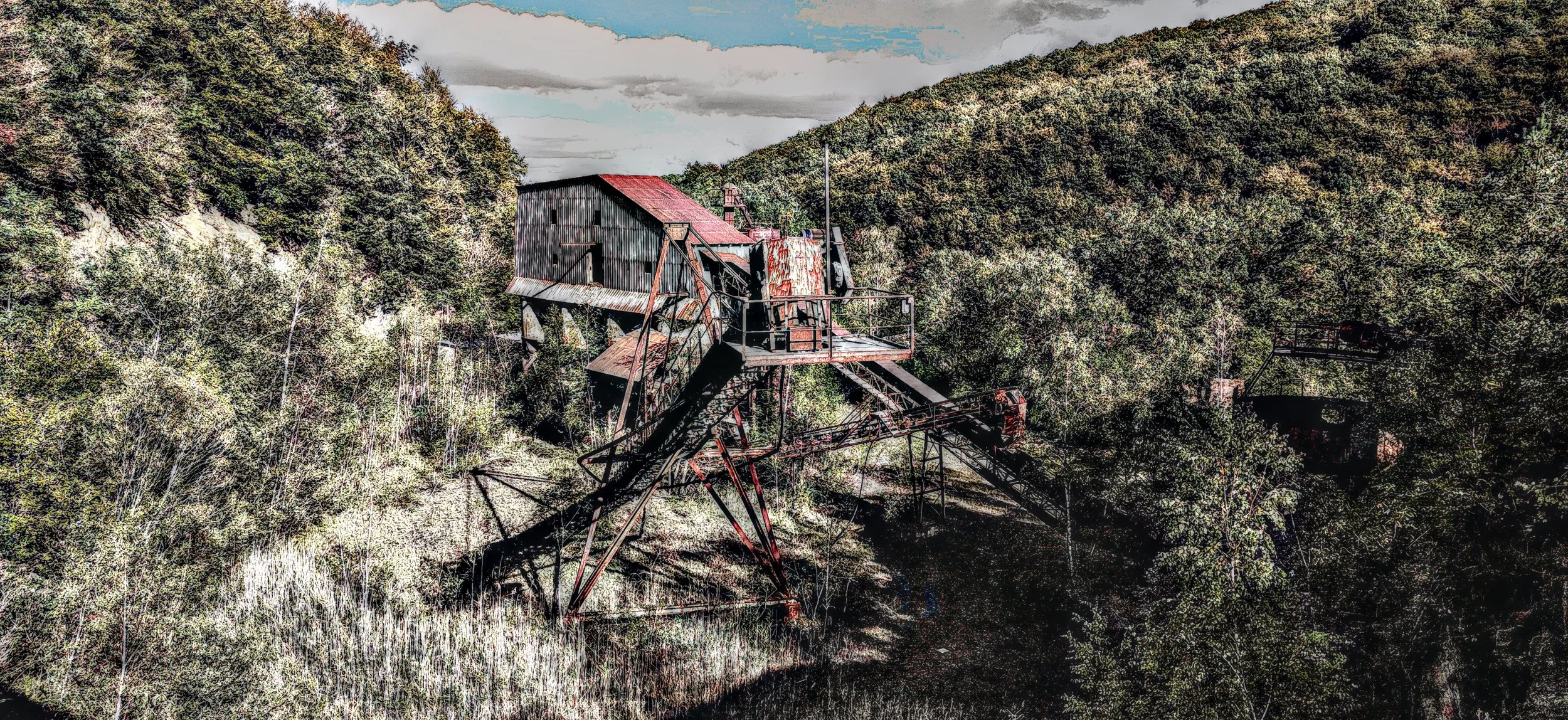 Verfallene alte Industrieanlage in einer bewaldeten Gegend, umgeben von Bäumen auf Hügeln, Wolken am Himmel