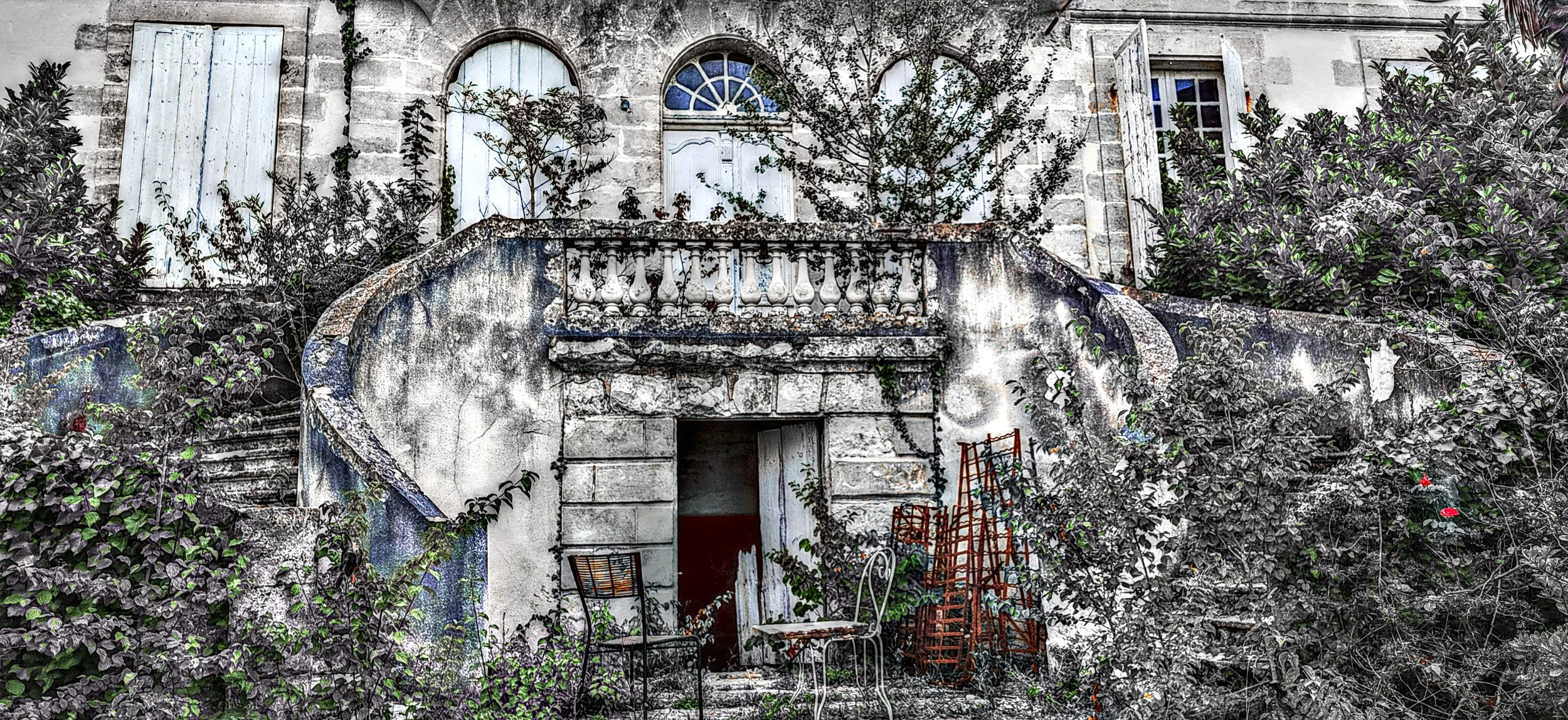 Verfallenes Gebäude mit einer aufwendigen Treppe und Balkon, überwuchert von Pflanzen und mit schließbaren Fenstern und Türen.