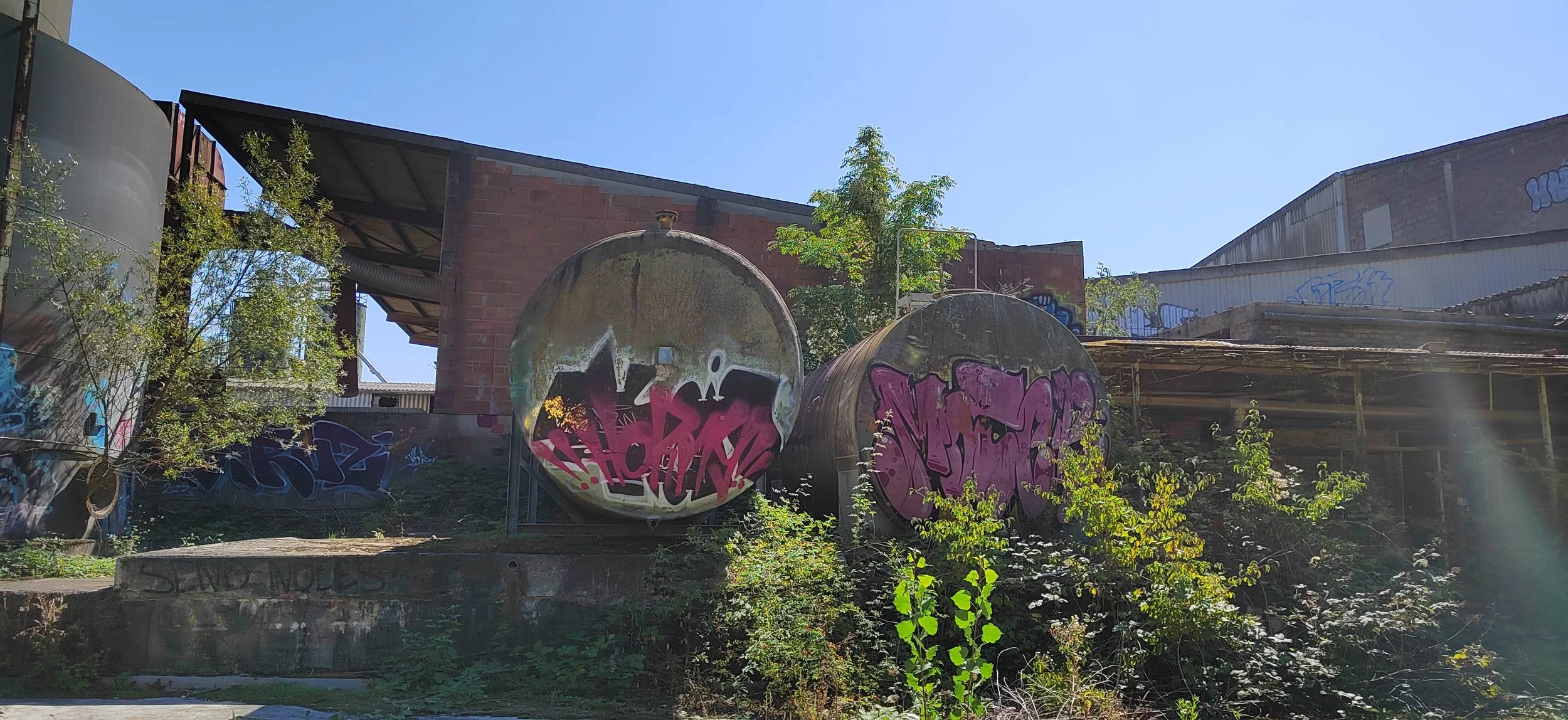 zwei alte, verrostete Tanks mit Graffiti in einer abbruchreifen Industrieanlage, umgeben von Pflanzen und Graffiti auf Mauern unter klarem Himmel