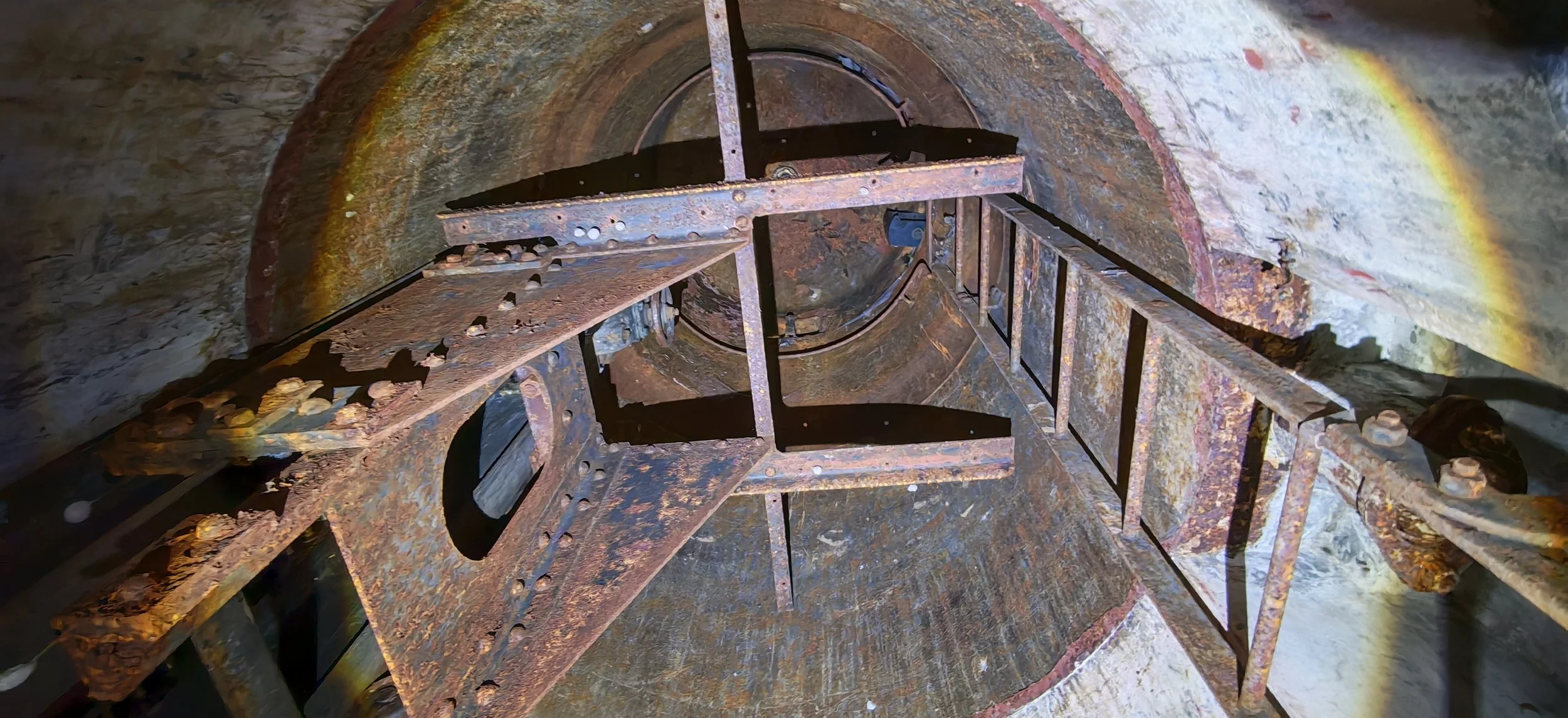 Blick in einen rostigen, engen Tunnel mit einer Metallleiter, die nach oben führt, und Mauern aus Stein.