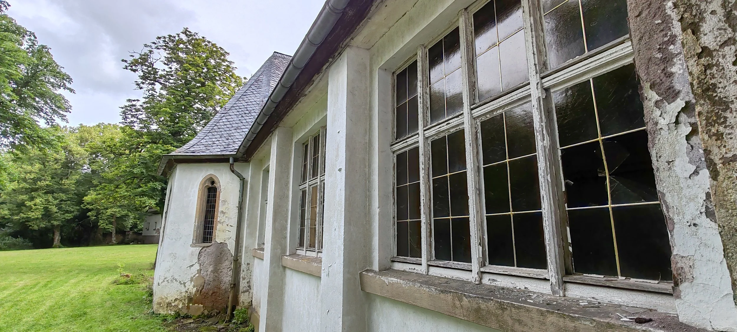 Alte verfallende Fenster an einer alten, verfallenden Steinmauer eines Gebäudes auf grünem Rasen vor Bäumen unter einem Himmel mit Wolken.