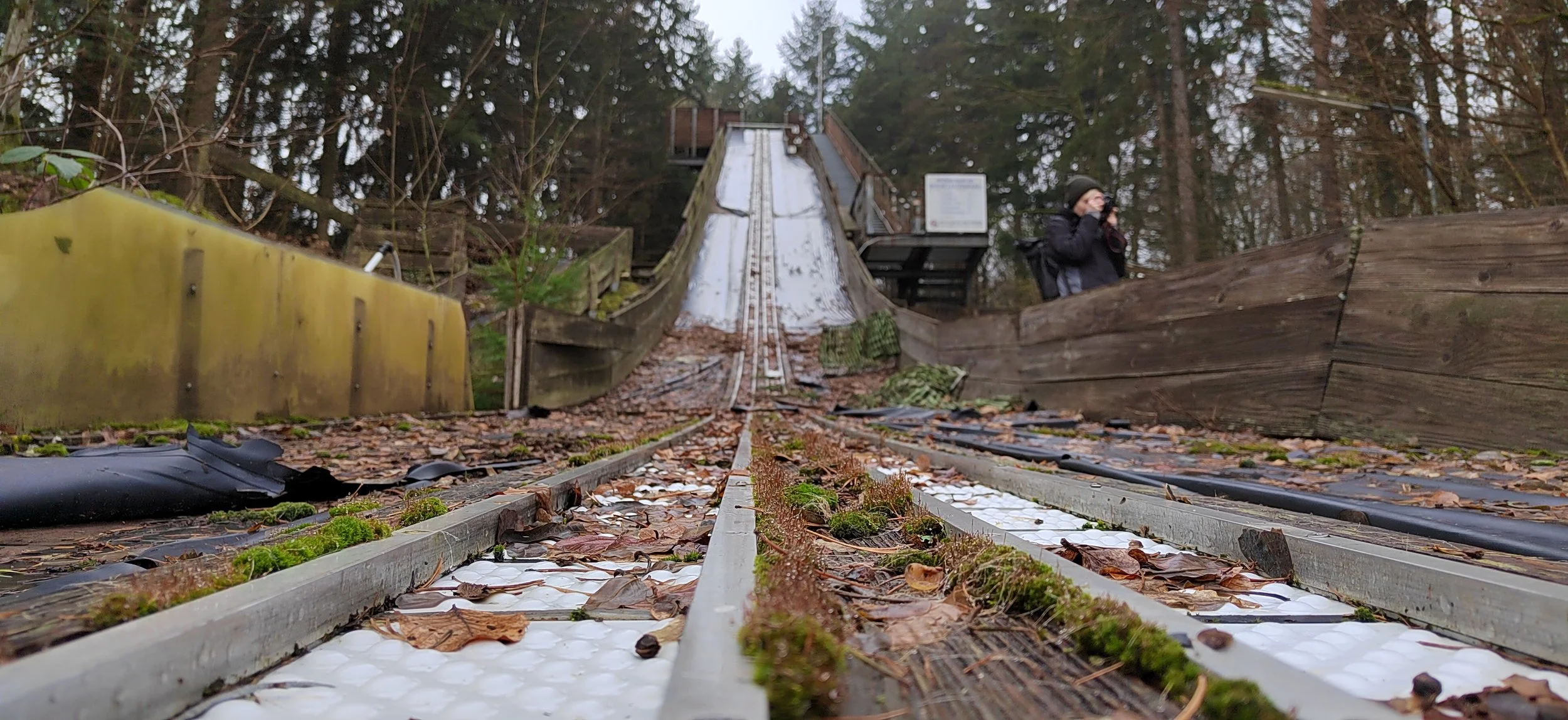 Blick entlang einer alten Rodelbahn im Wald, mit Moos und Laub bedeckt, im Hintergrund eine Person mit Kamera.