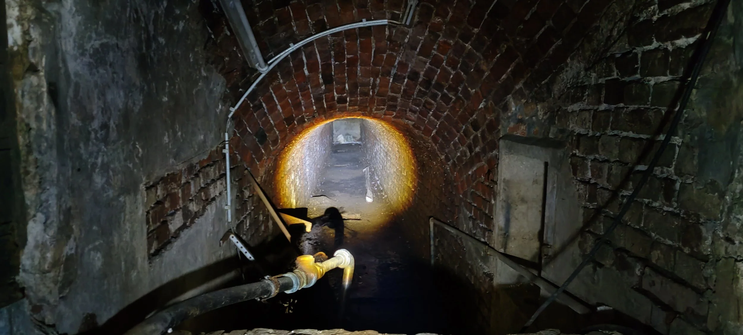 Ein Tunnel aus Ziegelsteinen, in dem eine Person arbeitet, Wasser auf dem Boden und Rohre an den Wänden.