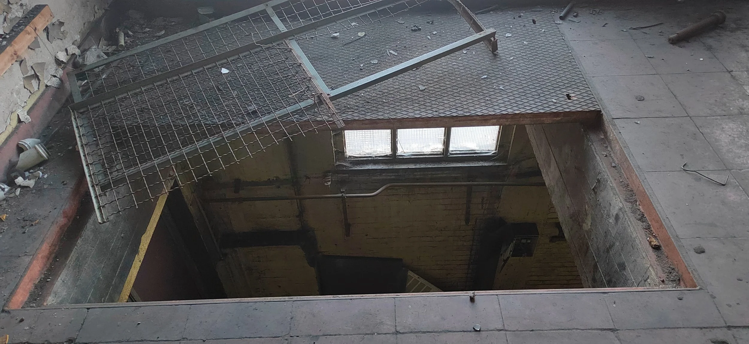 Blick in einen Baugrube mit Gitterrost und Steinen, Fenster im Hintergrund, und Baumaterialien auf dem Boden