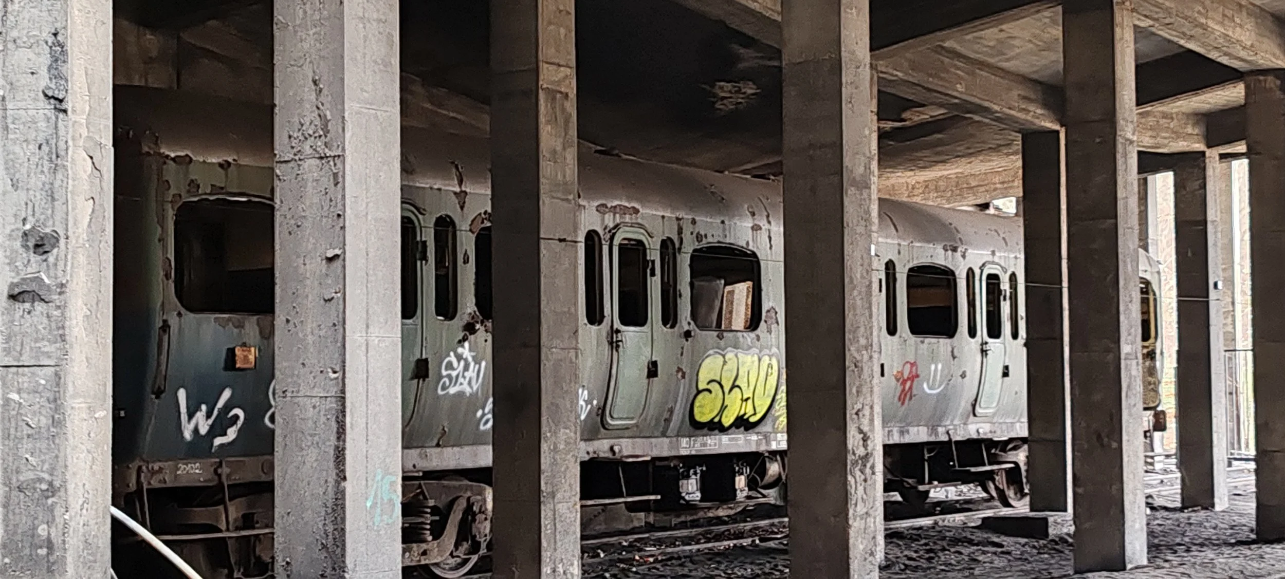 Verrosteter, verfallener Eisenbahnzug unter einer Brücke, umgeben von Betonstützen, mit Graffiti bemalt, Umgebung staubig und verfallen.