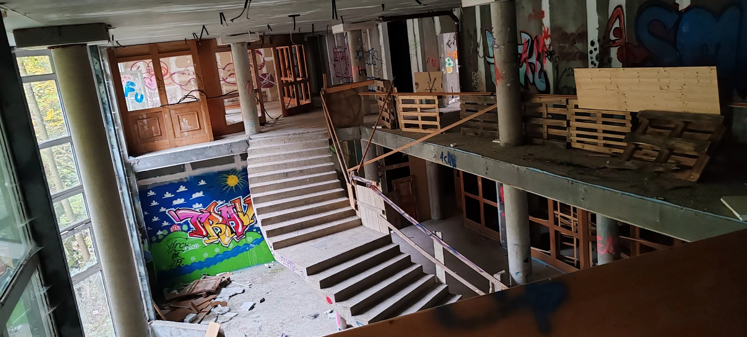 Ein mehrstöckiges Gebäude im Umbau mit einer Treppe, Graffiti an den Wänden, Bauholz und Baumaterialien, sowie offene Fenster und Türen.