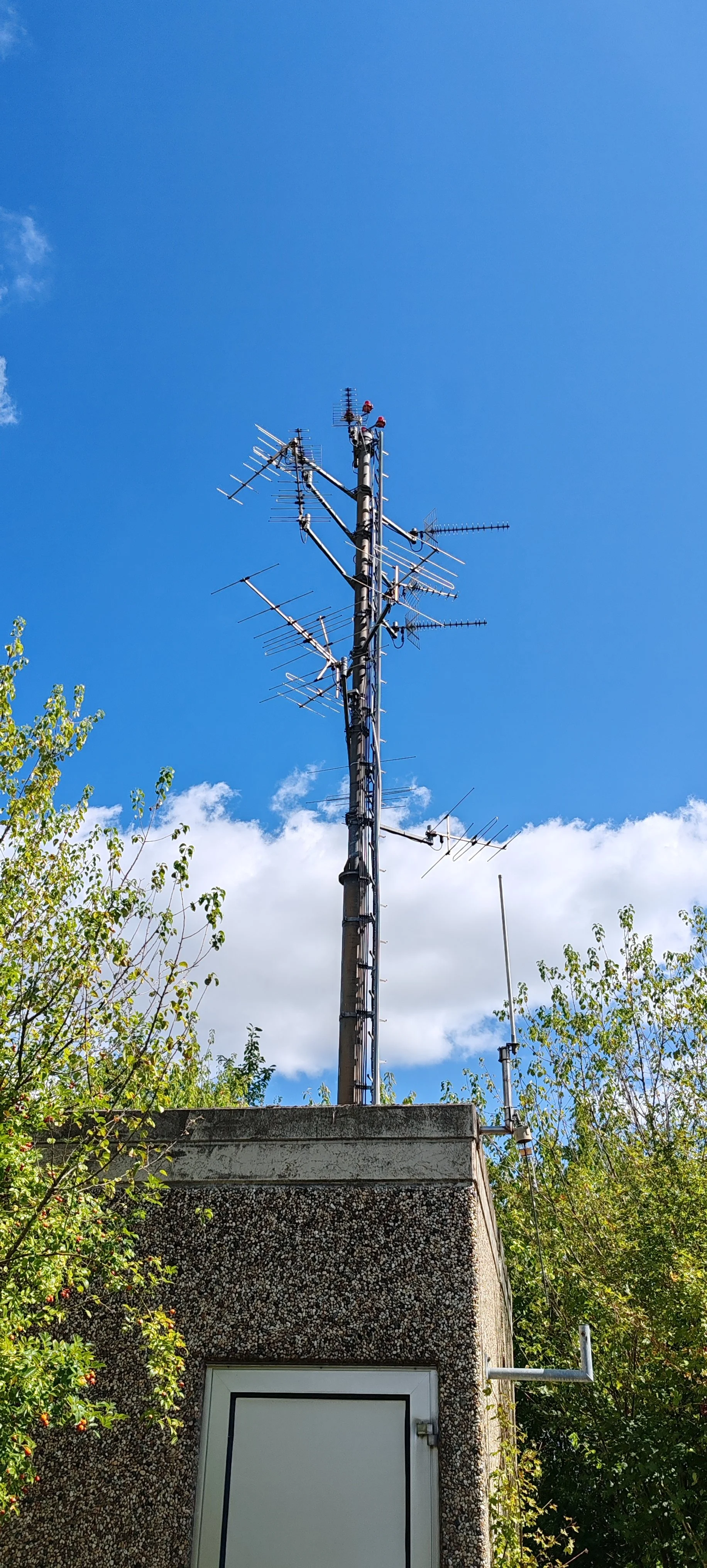 Ein Funkmast auf einem kleinen Gebäude im Freien, umgeben von Büschen und einem blauen Himmel mit wenigen Wolken.