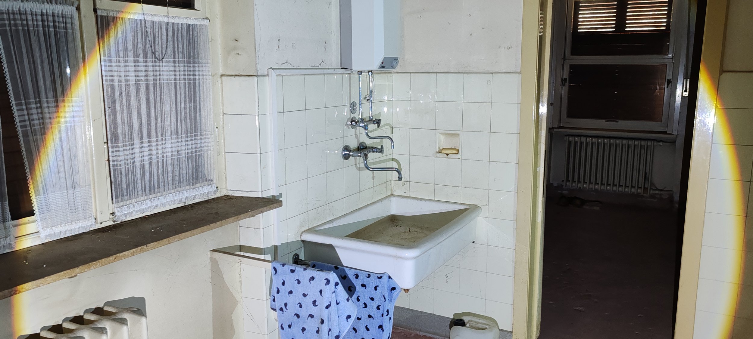 Altbau-Waschraum mit Waschbecken, Wasserhähnen und angebrachten Handtuch. Fenster mit weißen Gardinen, dunkler Raum im Hintergrund.