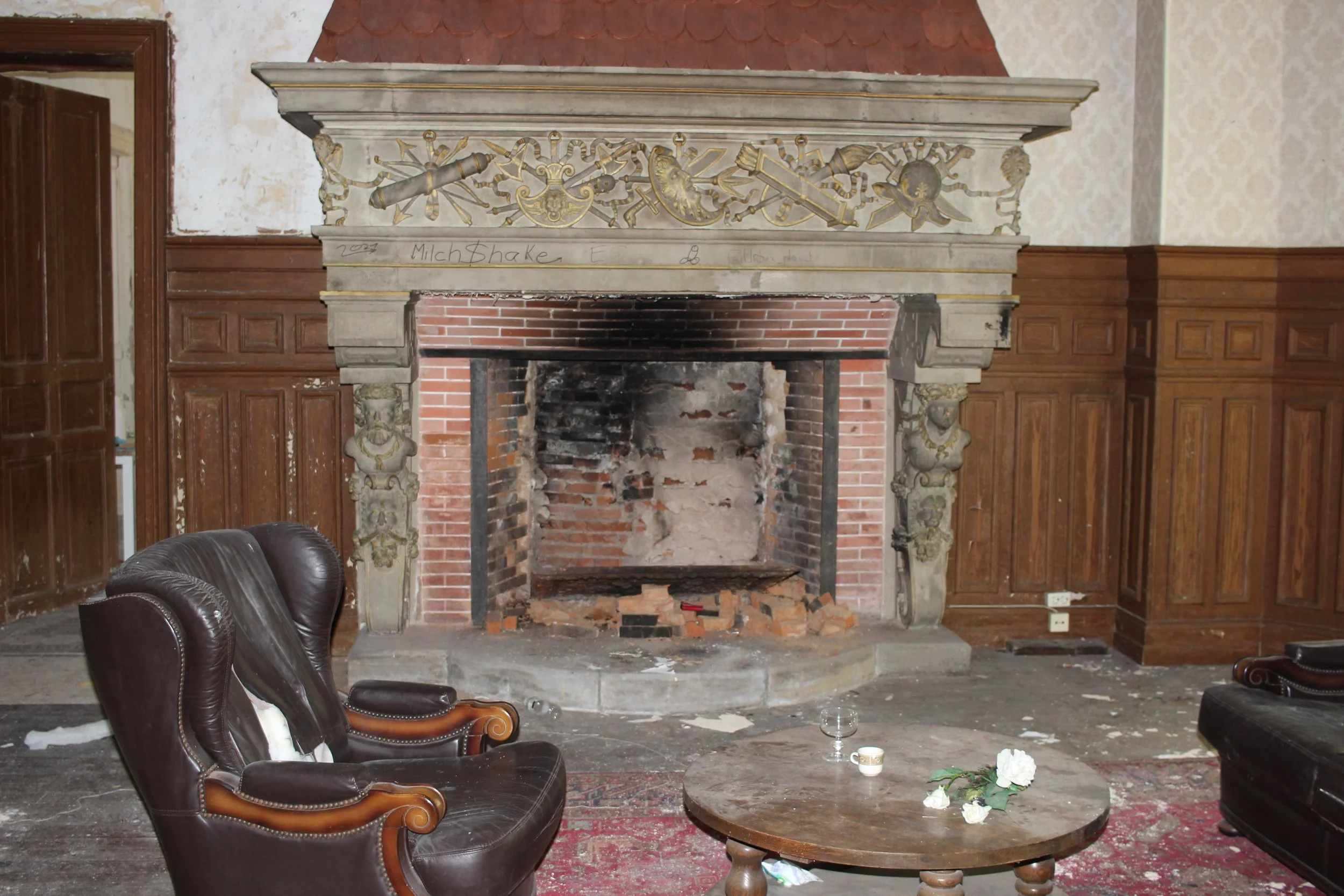 Ein alter, ungenutzter Kamin in einem Raum mit wetternder Wand und zerbrochenem Boden, umgeben von alten Ledersesseln und einem Holztisch, auf dem Blumen und eine Tasse stehen.