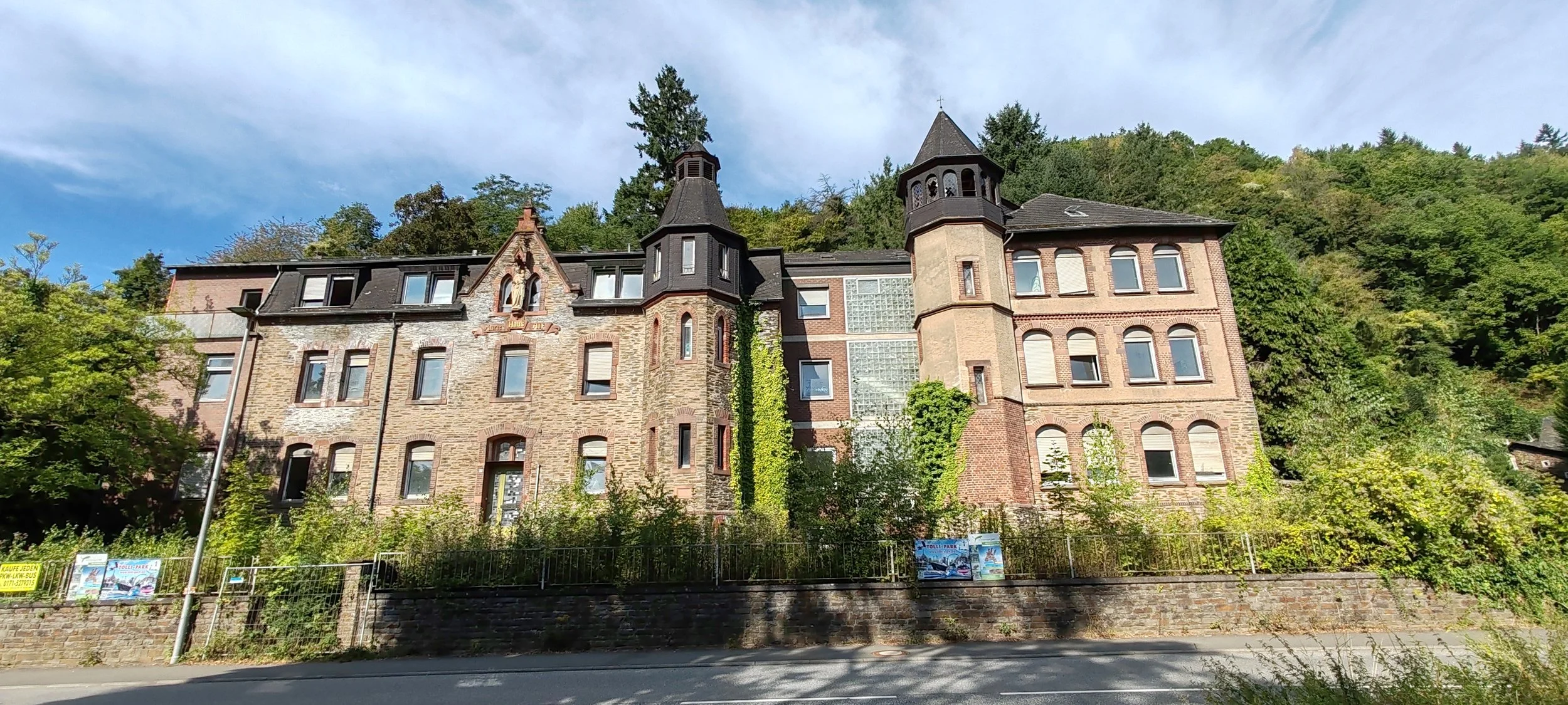 Historisches Gebäude mit Türmen und arched Fenstern, umgeben von Bäumen und einem Zaun, an einem sonnigen Tag.