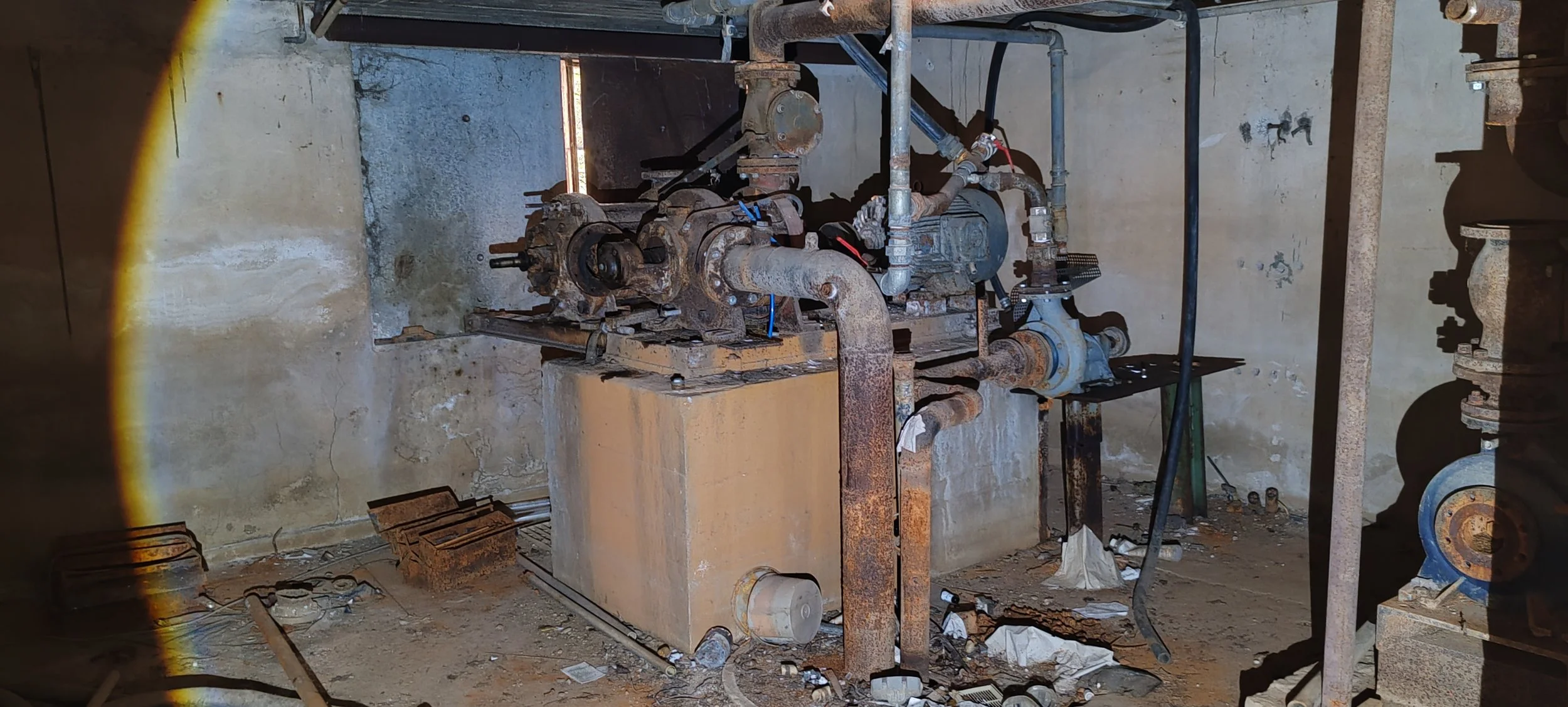 Alterte, rostige Maschinen mit Rohren und Pumpen in einem düsteren, ungepflegten Raum, vermutlich in einem Keller oder Technikraum.