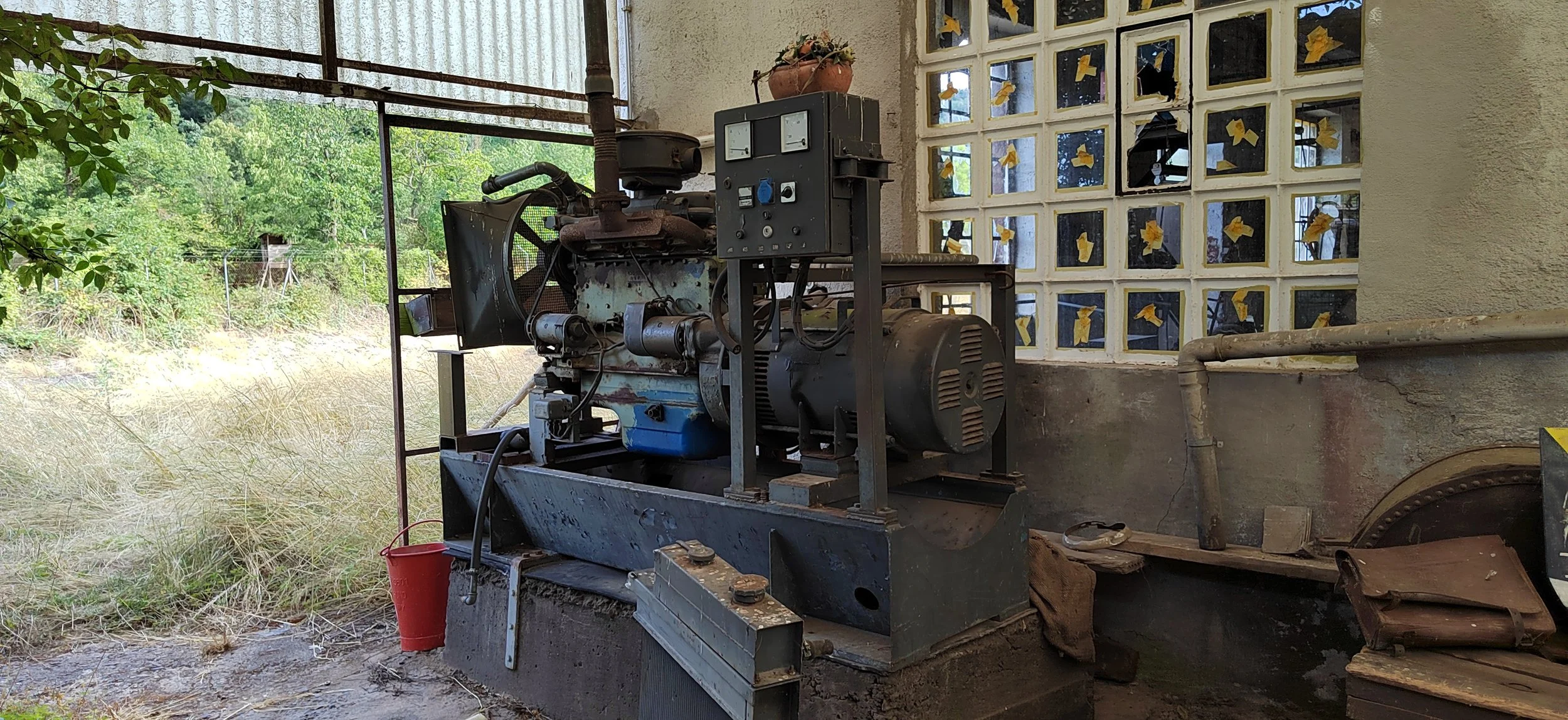 Alte, outdoor industrialische Maschine, die auf einem Betonsockel steht, mit einem weißen Mauerwerk im Hintergrund und einer Glasblockwand mit gelben Papierenten. Es gibt eine grüne Naturlandschaft im Hintergrund.