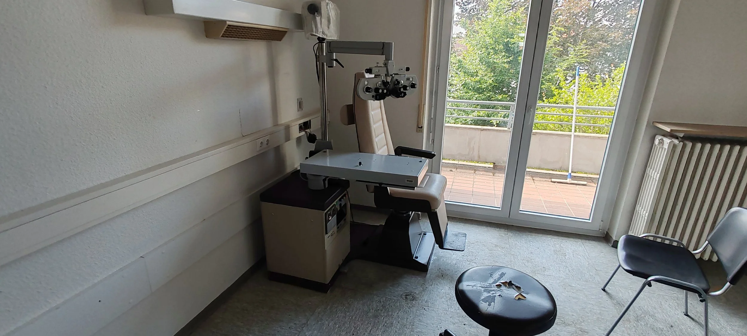 Ein Augenuntersuchungstisch mit Blick durch eine Glastür nach draußen auf eine Terrasse und Bäume, in einem Behandlungsraum.