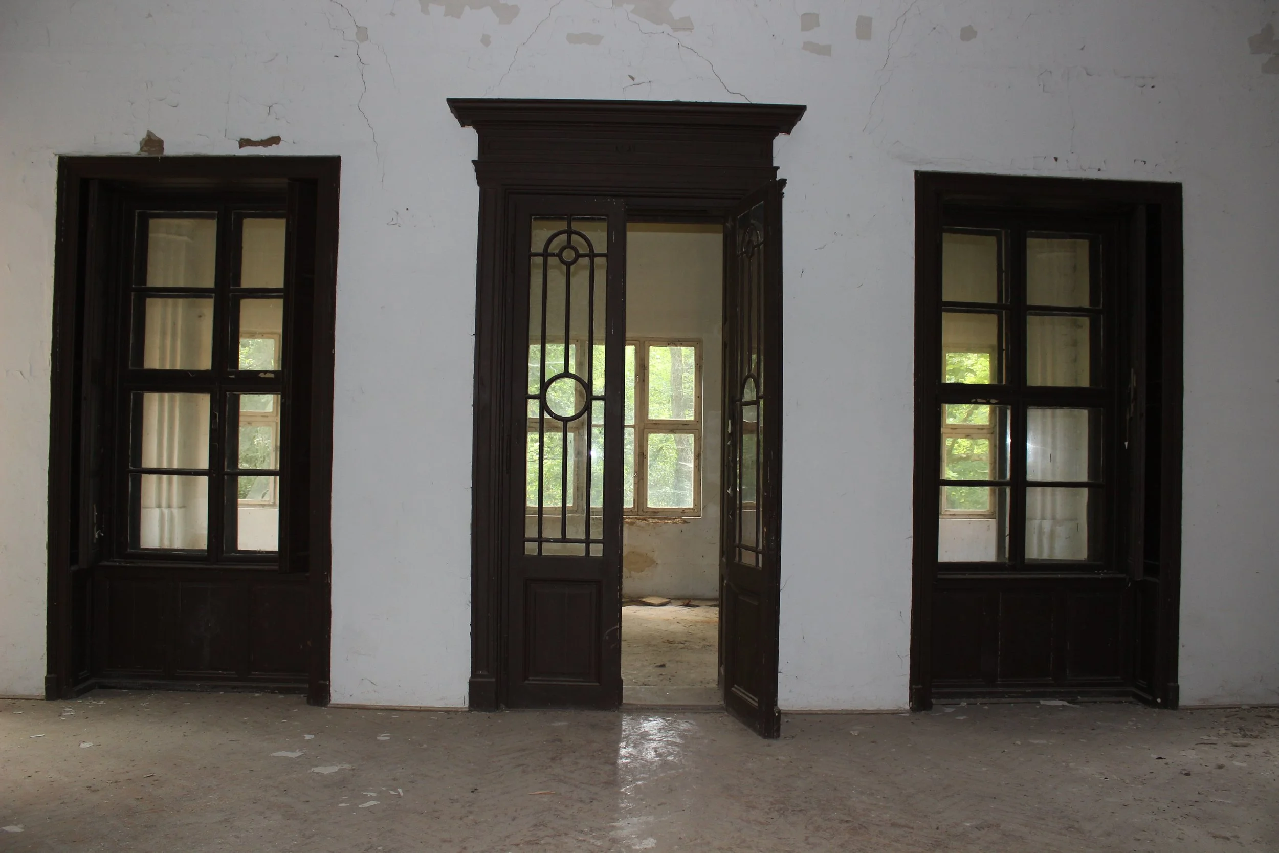 Innenansicht eines verfallenen Gebäudes mit drei Türen, die zu einem Raum mit großen Fenstern führen, dunkle Türen und zerbrochene Wände, Staub und Schmutz auf dem Boden.