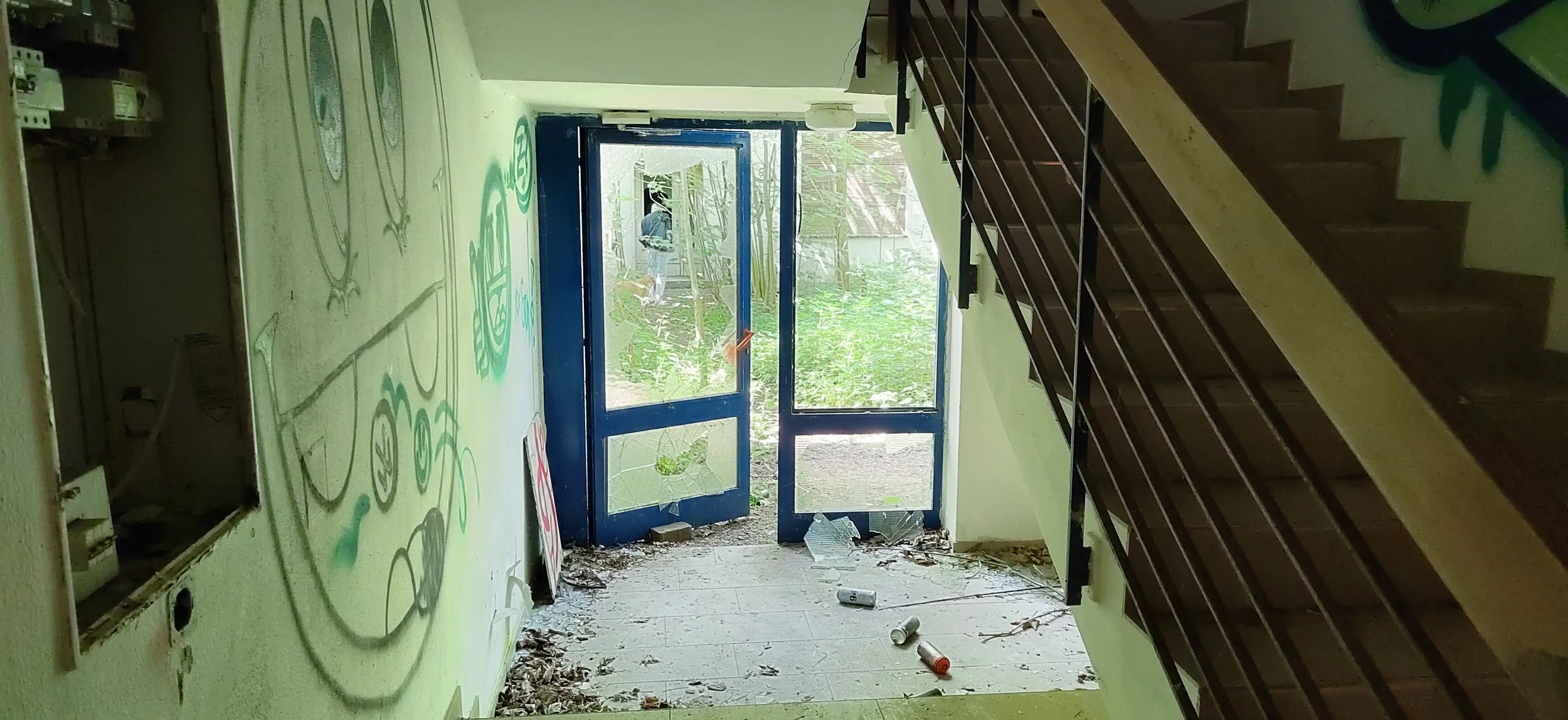 Innenansicht eines verlassenen Gebäudes mit Graffiti an der Wand und Müll auf dem Boden, blaue Tür zum Außengelände, Treppe auf der rechten Seite.