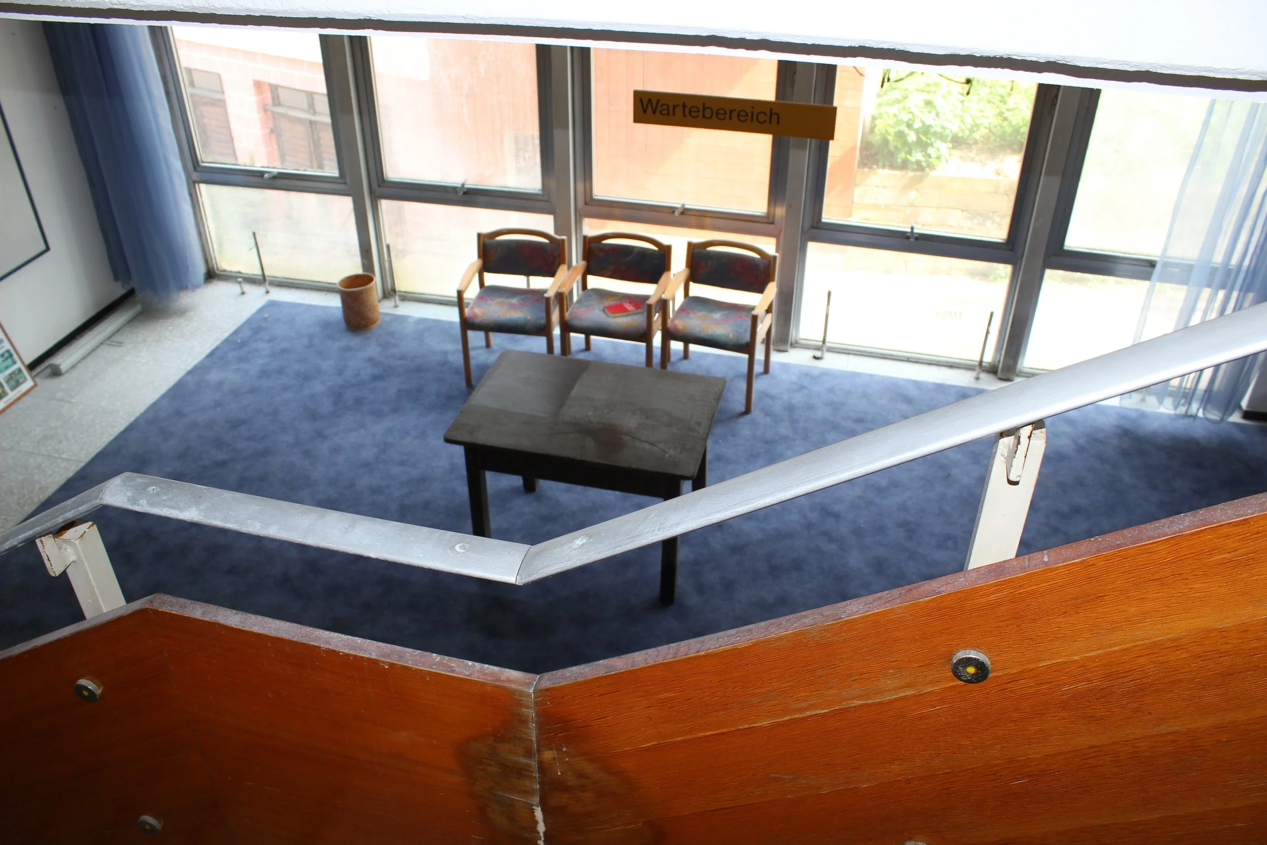 Warteschleife im Wartebereich mit drei Stühlen und einem kleinen Tisch vor großen Fenstern, die Tageslicht hereinlassen.