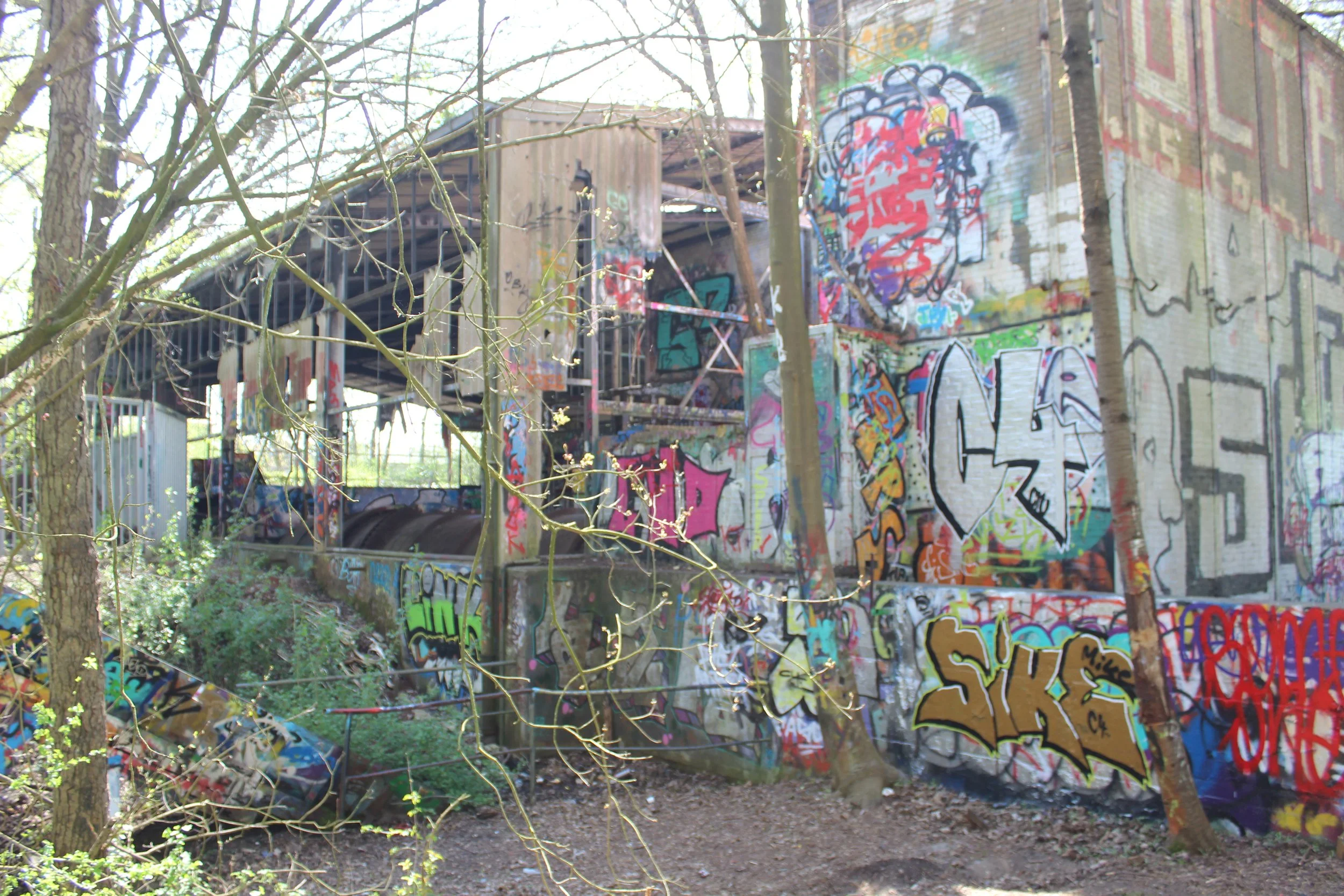 Verfallene Brücke und Mauern, bedeckt mit buntem Graffiti, in einem Park mit Bäumen und Pflanzen.