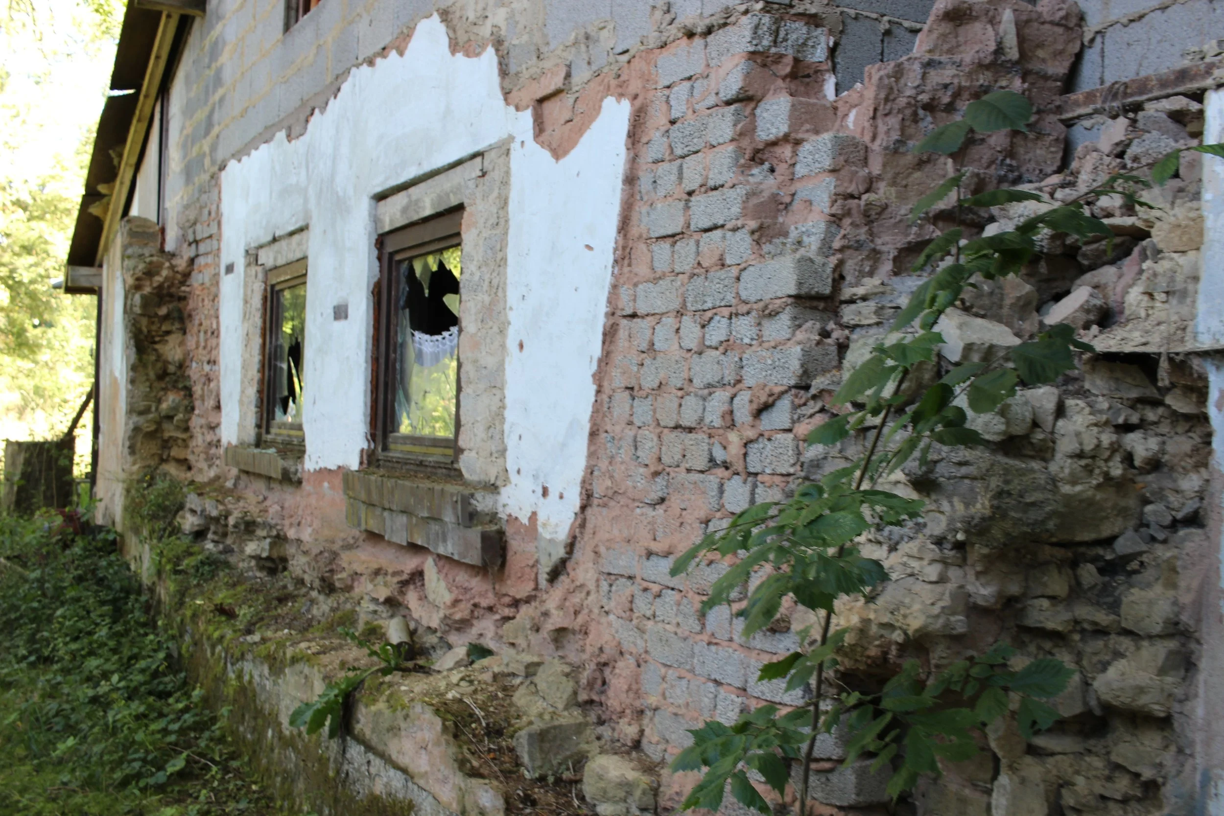 Eine alte, beschädigte Hauswand mit zerbrochenen Fenstern, teilweise verputzt und mit sichtbaren Ziegelmauern, umgeben von Grünpflanzen.