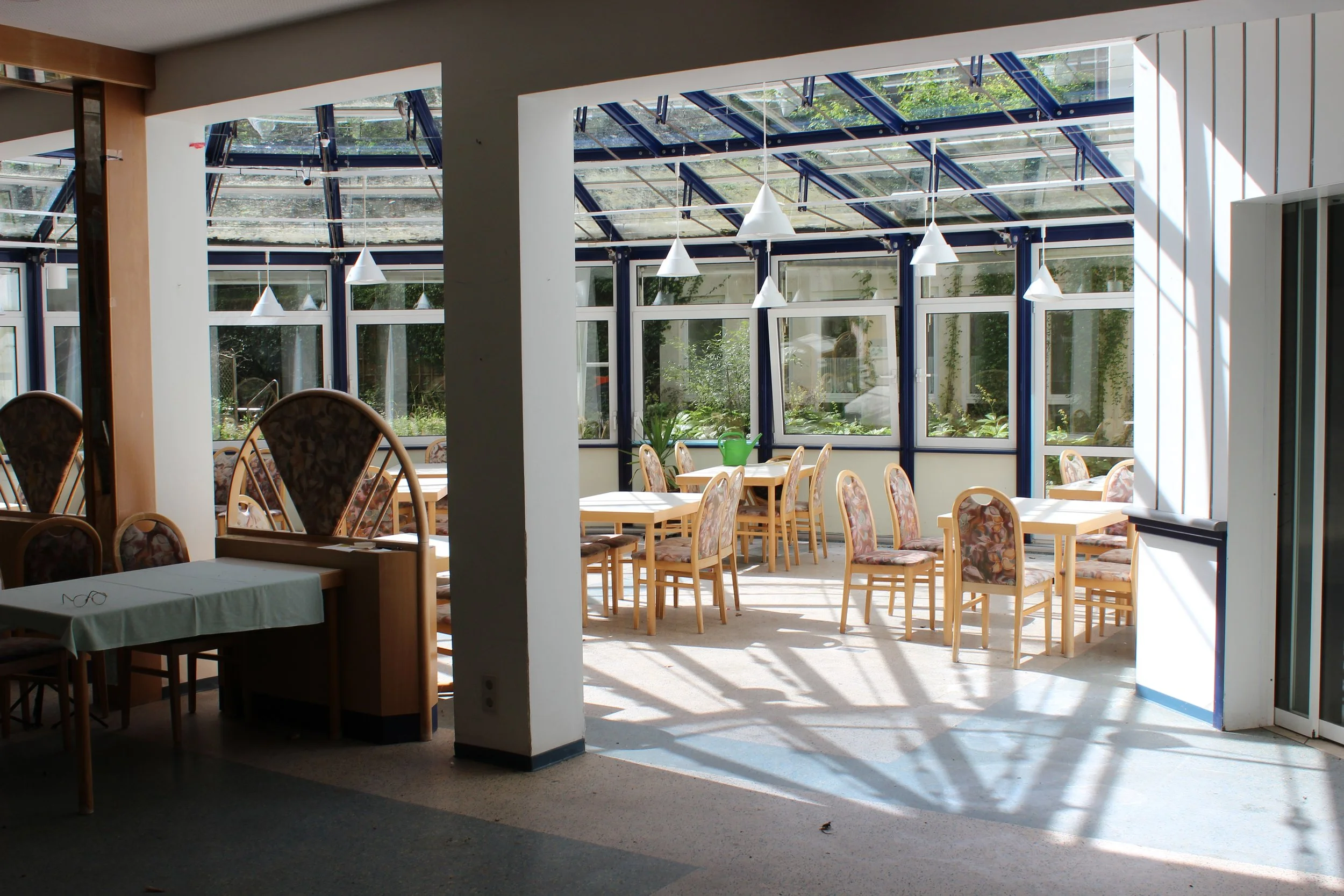 Innenraum eines Cafés oder Restaurants mit mehreren Tischen und Stühlen, viel Tageslicht durch große Fenster und eine gläserne Dachkonstruktion.