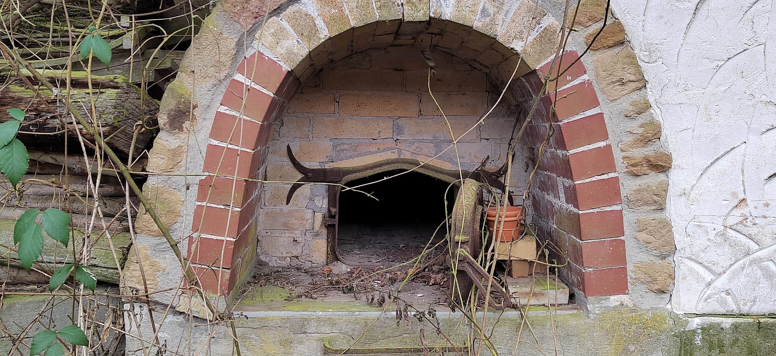 Ein verlassener Backofen aus Ziegeln und Steinen, mit eingewachsenem Grünzeug und einem Stück Holz davor.
