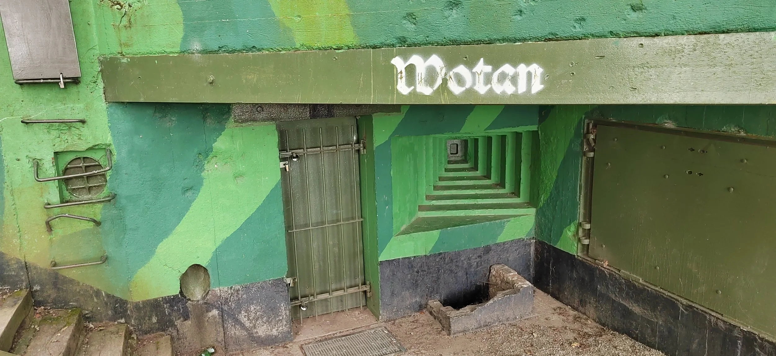Vertikale grüne Wand mit Metallgitter, Tür und Treppe, und der Schriftzug 'Moton' in weißer Schrift. Es sieht aus wie ein Eingang in eine unterirdische Passage, mit Treppen, die nach unten führen.