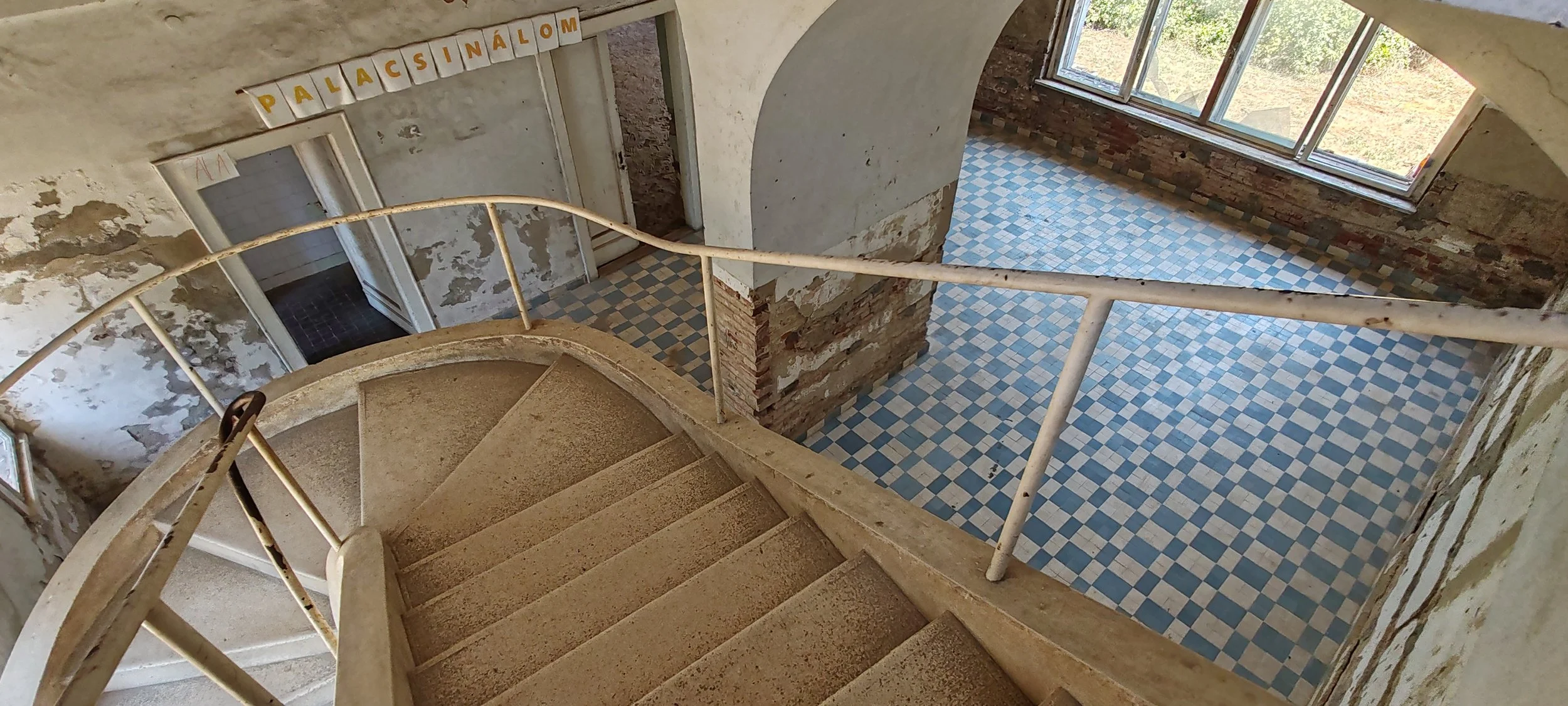 Innenansicht eines alten, vernachlässigten Gebäudes mit einer Spiraltreppe, abblätternder Wandfarbe und einem großen Fenster, durch das Tageslicht hereinfällt.